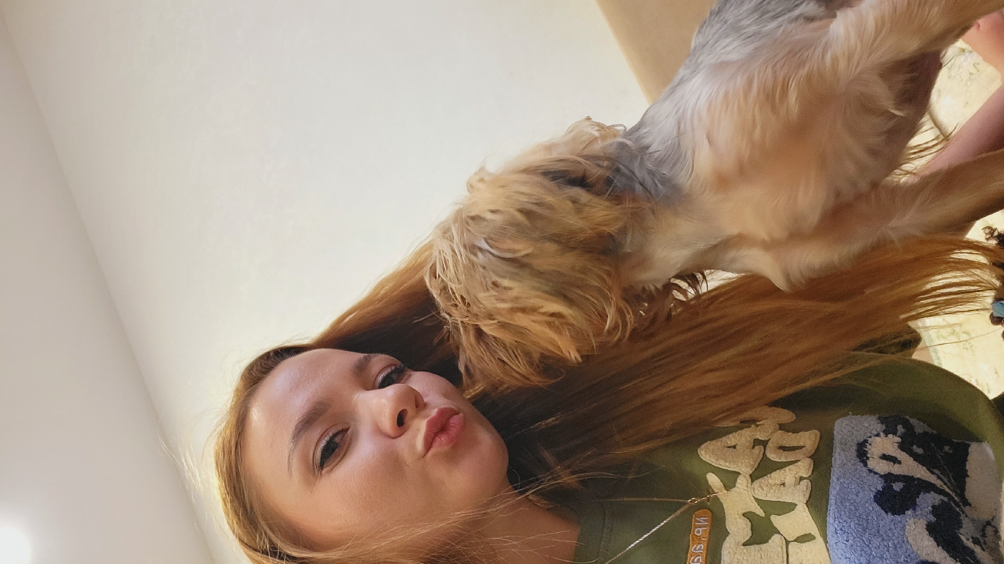 A close-up portrait captures a young woman and a small, shaggy-haired dog indoors in Chaikovsky, Russia. The image is taken from a slightly low angle, focusing on the subjects' faces and upper bodies.
The woman, on the left, has light skin, dark eyes, and long, straight auburn hair. She is making a 'kissing' face with puckered lips. She wears a green top featuring a large, textured design with white letters, possibly spelling "AD" or "BAD", and a blue pattern. A thin silver chain necklace with a small rectangular pendant, displaying the numbers "99", is visible around her neck.
The dog, positioned above and to the right of the woman, is a small breed with shaggy brown and grey fur. Its head, with longer, lighter brown hair, is turned towards the woman's head, appearing to sniff or nuzzle her hair. The dog's body is predominantly grey. The scene is brightly lit, indicating daytime. The background is a plain, light-colored wall or ceiling. The image conveys a sense of playfulness and affection between the woman and her pet.