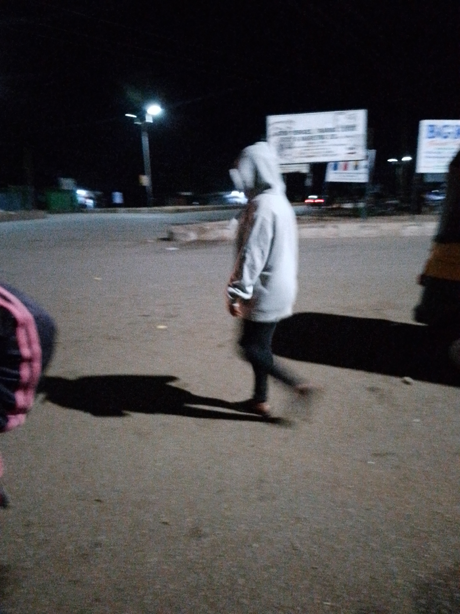 The image shows a street scene at night in Bukuru, Nigeria. The primary subject is a person wearing a grey hooded sweatshirt and dark pants, walking across a paved road. The person is in the middle of the frame and is partially blurred, suggesting motion. To the left of the frame, another person's legs and torso are visible, wearing a dark garment with pink stripes. To the right, another person is partially visible, wearing a yellow garment.
The setting appears to be an urban or semi-urban area with streetlights illuminating the scene. In the background, there are signs with text, though they are blurry and difficult to read. One sign in the center background appears to have multiple lines of text, possibly an advertisement or informational notice. Another sign to the right is also visible, with a portion of the word "BIG" clearly legible. There are also blurry lights from vehicles in the background, indicating traffic.
The time of day is clearly night, as evidenced by the darkness and the artificial lighting. The weather appears to be clear, with no signs of rain or adverse conditions. The overall mood is somewhat ambiguous due to the low light and blurriness, but the presence of people walking suggests normal evening activity. The ground is paved with asphalt and shows some signs of wear. Shadows are cast on the road by the individuals and any other objects.