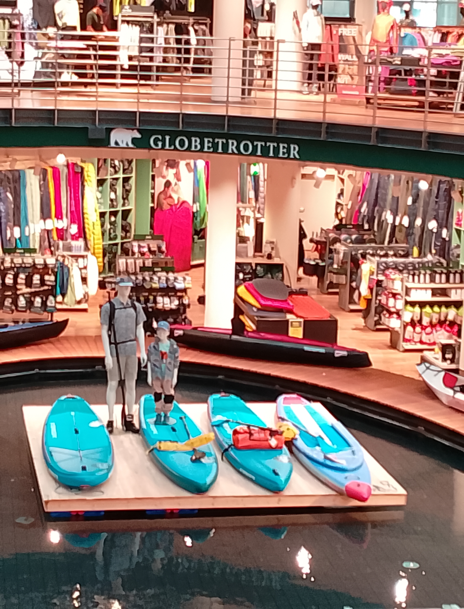 The image depicts an indoor retail environment, likely a sporting goods store, with the "GLOBETROTTER" logo visible. The scene showcases a display of kayaks on a wooden platform floating in a shallow body of water. Two mannequins, one adult-sized and one child-sized, are positioned next to the kayaks, dressed in outdoor apparel. The background reveals rows of shelves stocked with various clothing and outdoor equipment, typical of a retail setting. The lighting suggests an indoor, commercial space. The overall impression is of a product display designed to attract customers interested in water sports and outdoor activities.