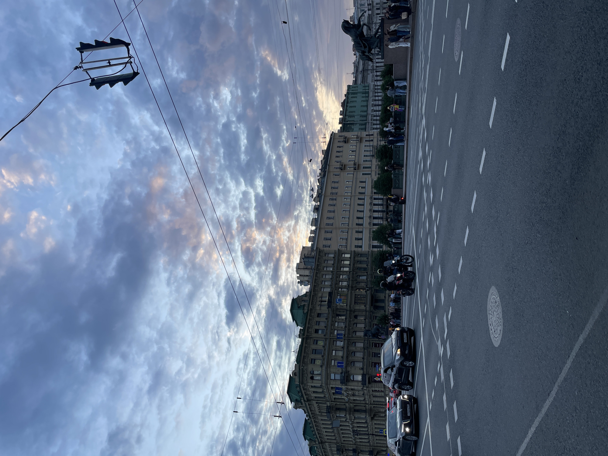 The image depicts a street scene in Saint Petersburg, Russia, likely on a bridge or a wide avenue, given the presence of multiple lanes of traffic and buildings lining the thoroughfare. The time of day appears to be late afternoon or early evening, as indicated by the dramatic, cloud-filled sky with soft, warm light breaking through in the distance.
In the foreground, a paved road with clear lane markings and a manhole cover is visible. Several vehicles are in motion or stopped: a black SUV, a black sedan with its headlights on, and two motorcycles carrying riders.
In the middle ground, a group of pedestrians is gathered on a sidewalk or promenade behind a decorative railing. Further back, a line of multi-story buildings with traditional architecture and green roofs dominates the skyline. The buildings exhibit varied architectural styles, suggesting a historical urban environment. To the right, a large bronze sculpture of a horse and rider is positioned on a pedestal.
Suspended above the roadway are traffic lights and a network of power or communication lines, typical of an urban setting. The overall impression is of a bustling city street at dusk, with a blend of modern vehicles and historical architecture under a dynamic sky.