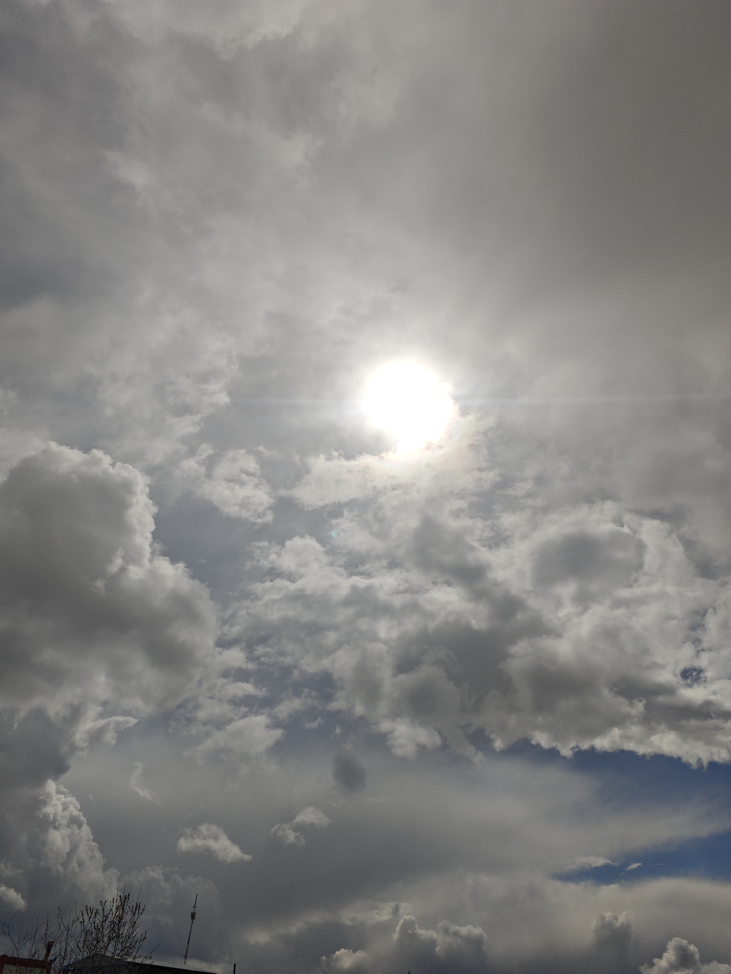 The image shows a sky filled with clouds of varying shades of gray and white, with the sun shining brightly through a break in the cloud cover. In the lower portion of the image, there are hints of buildings and vegetation, suggesting an urban location. The photograph was taken in Unknown, Russia.