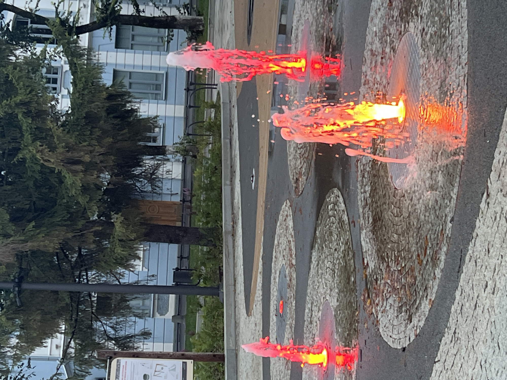 An outdoor shot shows a decorative fountain with jets of water illuminated by red and orange lights. The fountain is situated in a paved area with concentric circular patterns of small stones. In the background, a light blue building with white trim and dark windows stands behind a line of trees. A lamppost is visible to the left of the building, and a trash can is positioned near it. Several trees with dark green foliage are in front of the building.