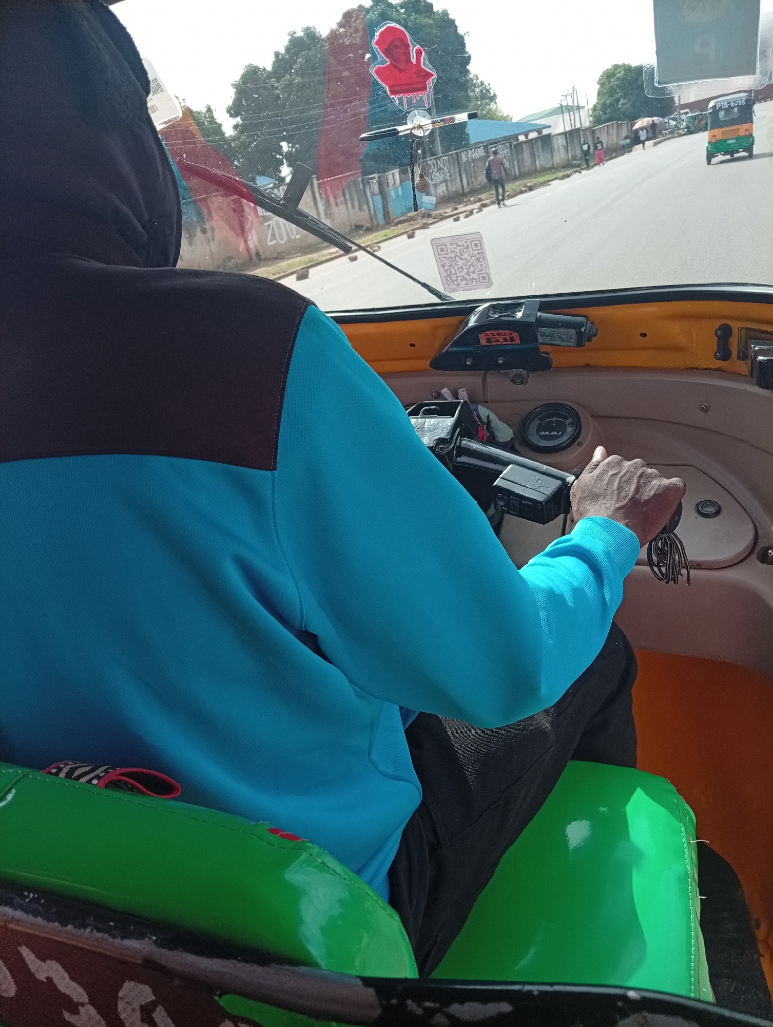 A driver, seen from the rear, operates a "keke" (auto-rickshaw) on a road in Jos, Nigeria. The driver wears a black hoodie and a blue top. His right hand is on the handlebar. The "keke" has a yellow dashboard and a green seat. The windshield of the "keke" has a QR code sticker and a small red figure hanging from it. Through the windshield, a road with pedestrians and another "keke" can be seen. Buildings and trees are visible on the side of the road.