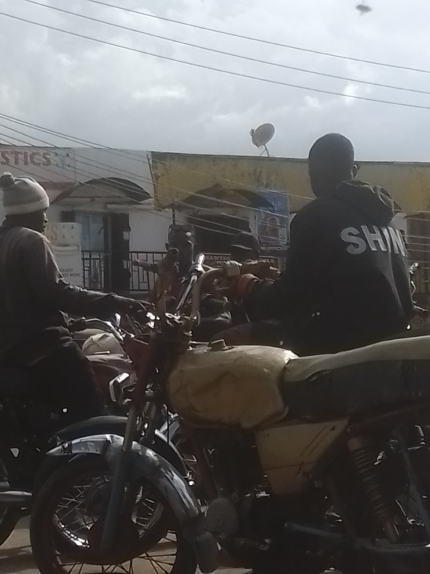 The image depicts a street scene with several motorcycles and people. In the foreground, a motorcycle with a faded yellow fuel tank and black seat is partially visible. Behind it, another motorcycle, mostly obscured, has a dark red fuel tank.
Two individuals are seated on motorcycles. The person on the left wears a grey hooded sweatshirt and a light grey beanie with a pom-pom. They are looking to their left. The person on the right is wearing a black hooded sweatshirt with the word "SHIN" printed in large white letters across the back. They are also seated on a motorcycle.
In the background, buildings line the street. One building has a white facade with text that is partially visible, including "STICS". Another building features a yellow awning. A satellite dish is visible on top of a structure in the background. Power lines are strung across the sky.
The weather appears overcast, with a cloudy sky. The lighting suggests it is daytime, possibly late morning or afternoon. The scene has a casual, everyday feel, likely a street in a populated area. No specific event or emotional expression is clearly conveyed.