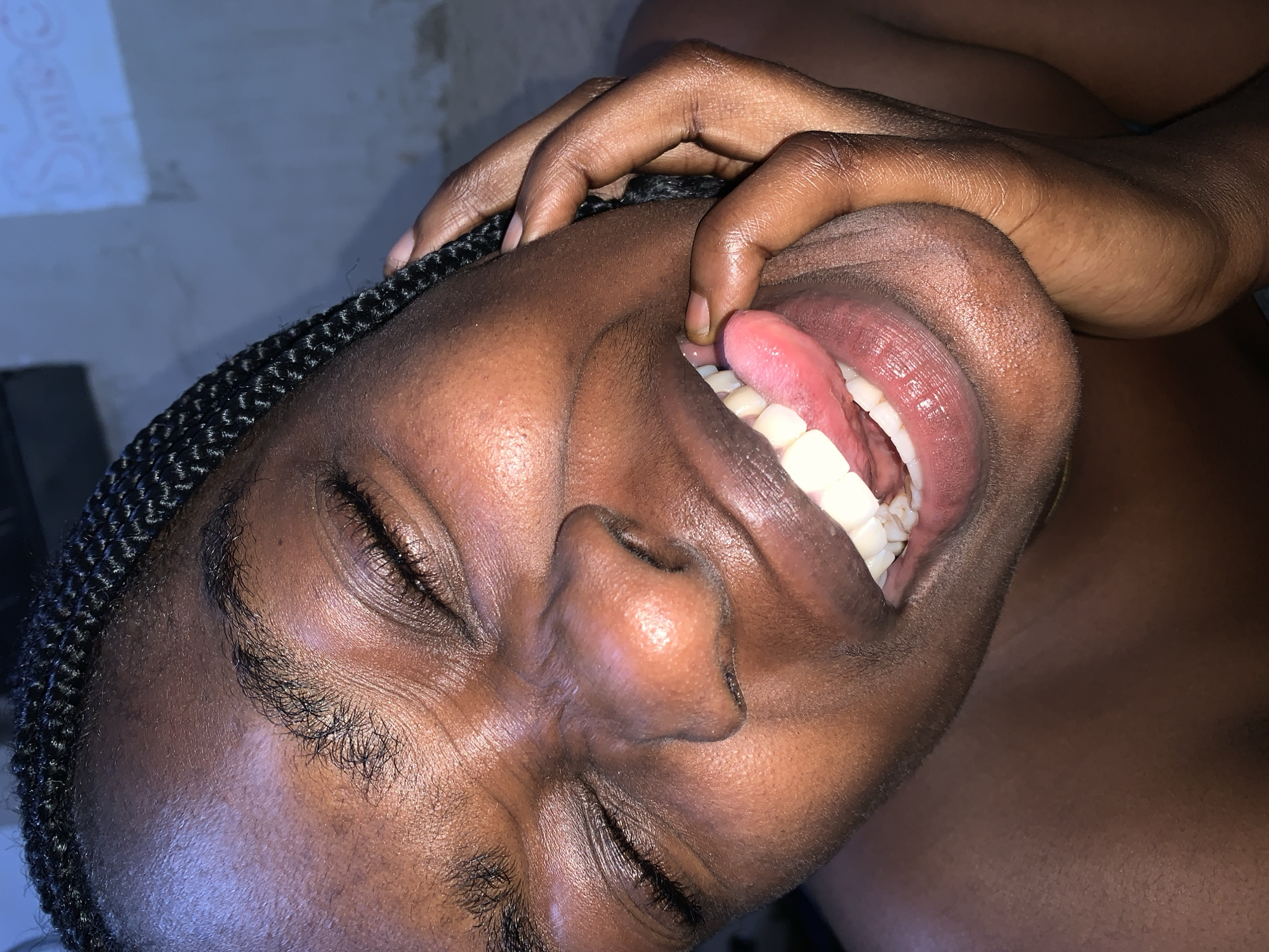 A close-up, high-angle shot shows a Black person with their eyes closed and a wide, playful grimace, with their tongue sticking out and touching the side of their face. Their hand is placed on their cheek, and their dark braided hair frames their face. The background is a blurred wall with a white chalk drawing of the word "SMILE." The lighting is dim, casting shadows and highlighting the person's skin. This image was taken in Rafin Par, Nigeria.