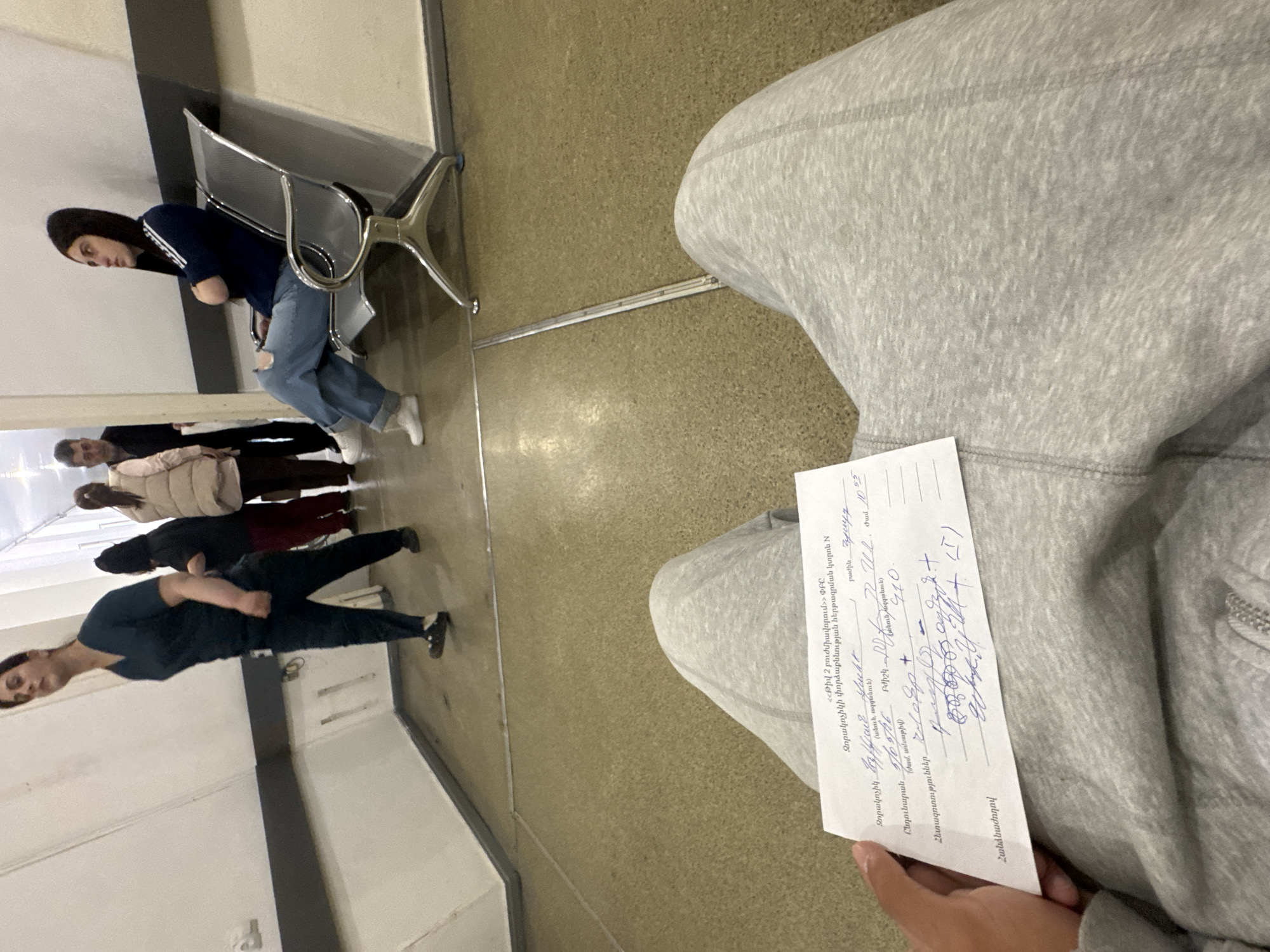 The image shows a person seated in a waiting area, possibly in a clinic or hospital, as indicated by the "Թիվ 2 բուժմիավորում" inscription on the form in their hand. The person wears grey sweatpants. The form appears to be a medical document, possibly related to a checkup for military conscription, as the text includes the word "Զորակոչիկի" which translates to "conscript" in English. Several other people are waiting in the same area. One is sitting on a metal chair. The location may be in Yerevan, Armenia, based on the text language.