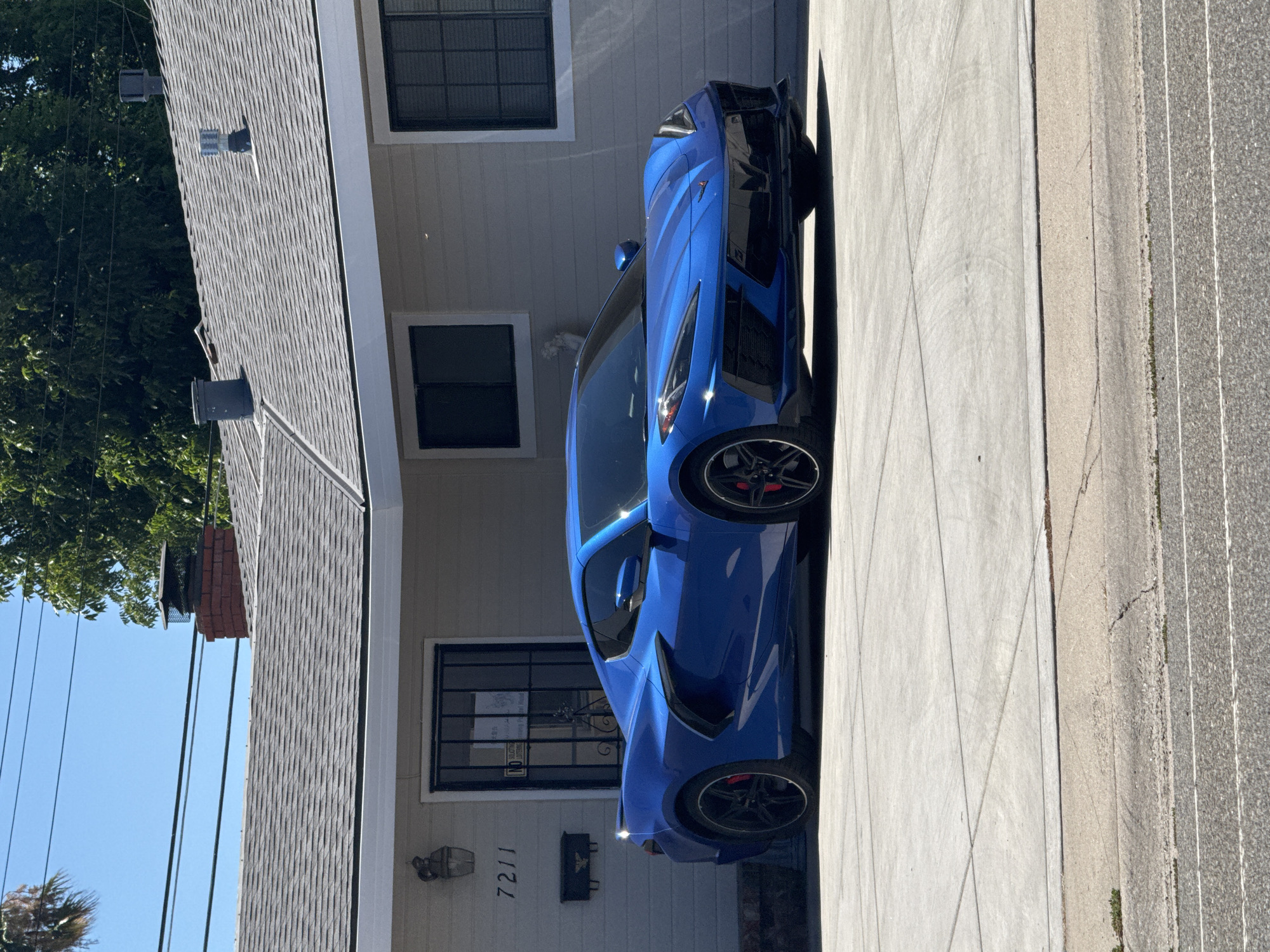 A bright blue Chevrolet Corvette C8 is parked on a concrete driveway in front of a single-story house in Citrus Heights, California. The house has a gray exterior with white trim and a gray shingled roof. There are two windows visible on the front of the house, one to the left of the entryway and another to the right. The house number "7211" is visible to the left of the entryway. The car is angled towards the viewer, with its front end slightly to the right. The sky is clear and blue, with some green trees visible behind the house. Power lines are strung across the sky.
