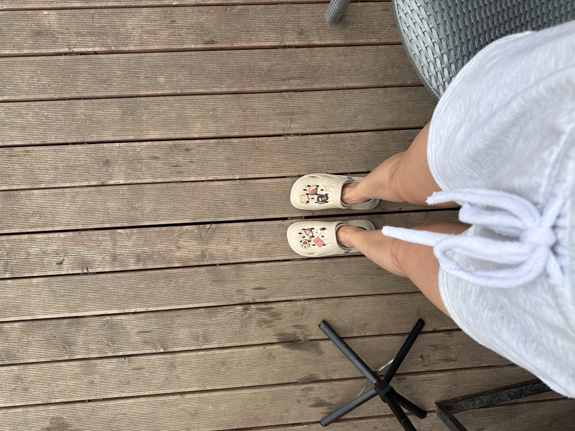 This is a high-angle, full-body shot of a person standing on a wooden deck. The person is wearing light-colored Crocs with cartoon character charms and a white drawstring dress. To the left of the person is a black tripod stand. To the right, a portion of a gray woven outdoor chair is visible. The wooden deck appears weathered and is composed of planks running vertically in the frame. The scene is outdoors, likely in a residential or recreational area. There is no discernible information in the image to indicate a specific location in the city of Semiluki, Russia.