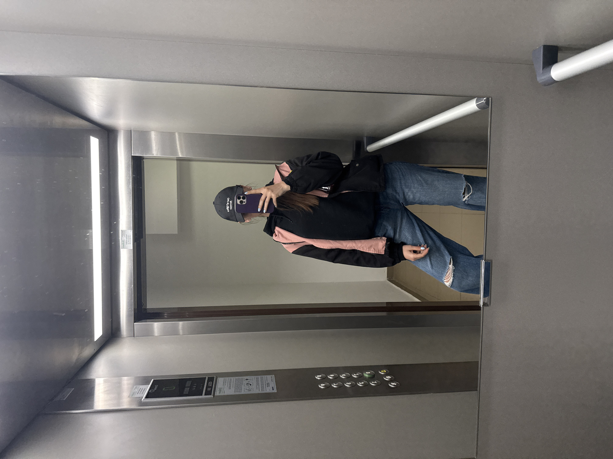 A person stands in an elevator, visible as a reflection in the mirror. They are wearing a dark baseball cap, a black jacket with pink accents, and ripped blue jeans. They are holding a phone up to their face, seemingly taking a photo. The elevator interior is simple, with stainless steel walls, buttons, and a handrail. The reflection shows a hallway outside the elevator door. The image was taken in an elevator in Plovdiv, Bulgaria.