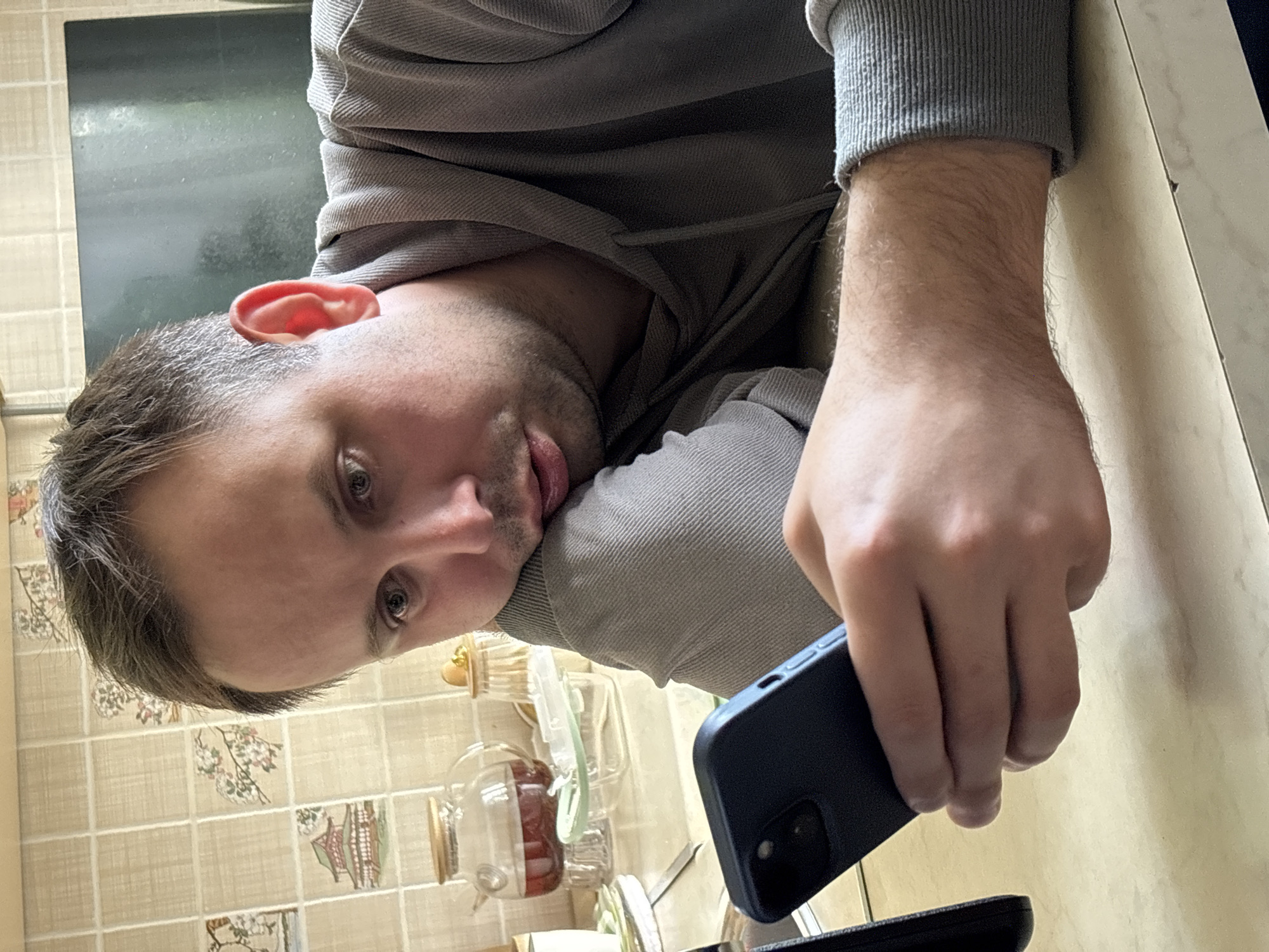 A man with short, light-brown hair, wearing a gray hoodie, is holding a dark-blue smartphone. He is indoors, likely in a kitchen or bathroom, given the tiled wall with a floral and oriental design in the background, as well as various glass containers and utensils on the surface. The location is not Rusanovo, Russia.