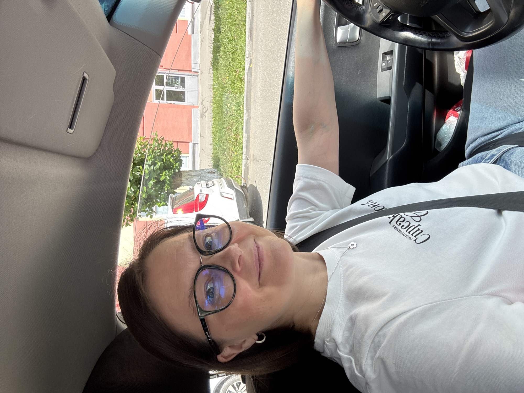 A woman with dark brown hair and glasses is sitting in the driver's seat of a car, looking at the camera. She is wearing a white t-shirt with a graphic print that says "Cupcake Corner" and a silver necklace with a flower pendant. Her right arm is resting on the car door. Outside the car window, a building with a red facade and white doors is visible, along with trees and a street. The image appears to be taken in the city of Izhevsk, Russia, based on external context.