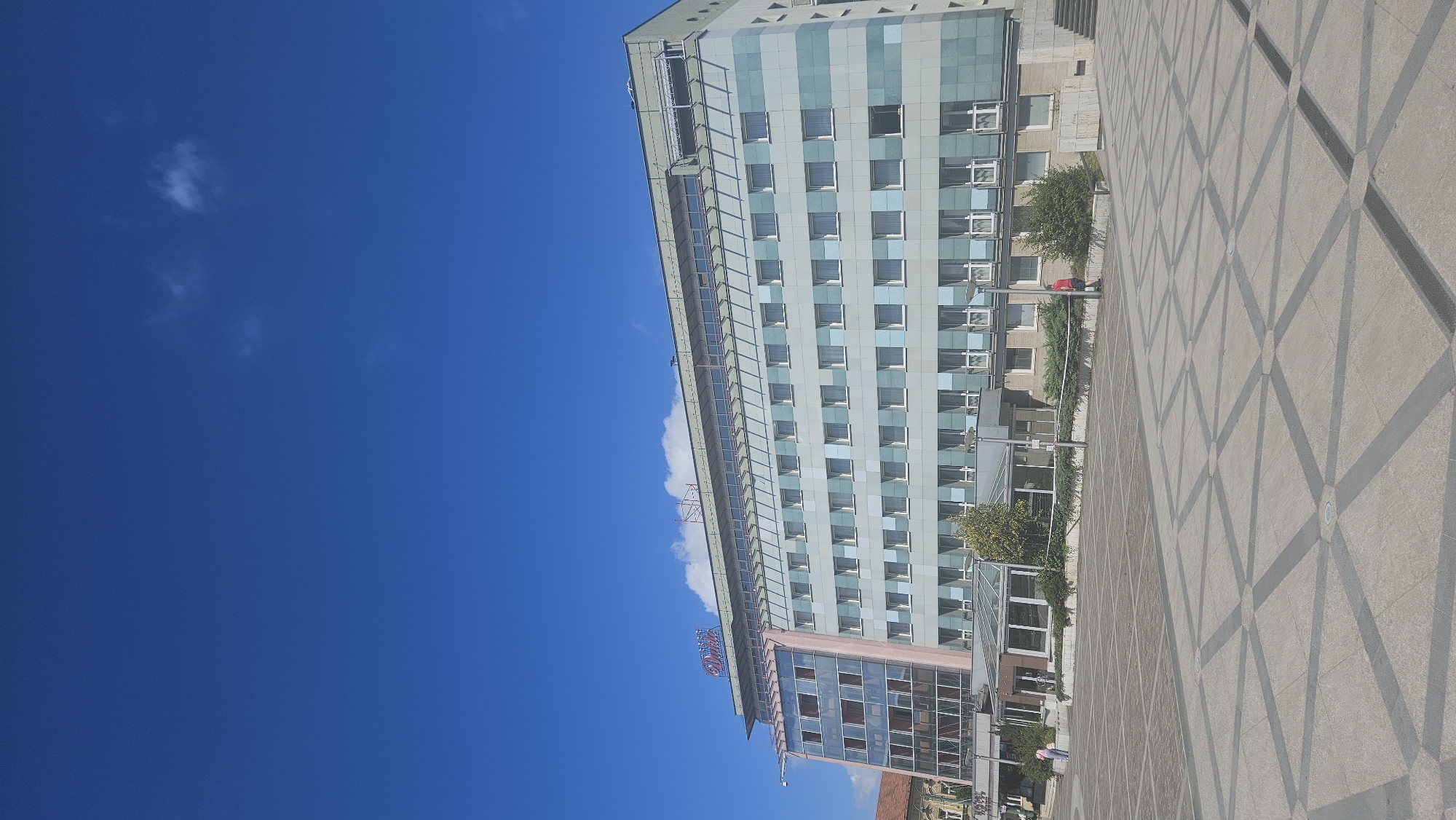 The image shows a modern multi-story hotel building with a distinctive facade featuring rows of windows. The building has a large glass-covered entrance area at the base and a prominent sign on the roof that reads "HOTEL SLOVAN". The hotel is situated in an open, paved plaza with geometric patterns on the ground. A few people are visible in the plaza. To the left of the hotel, part of another building with a reddish roof is visible. The sky above is a clear, bright blue with a few wispy clouds. The scene depicts the Hotel Slávia in the city of Prešov, Slovakia.