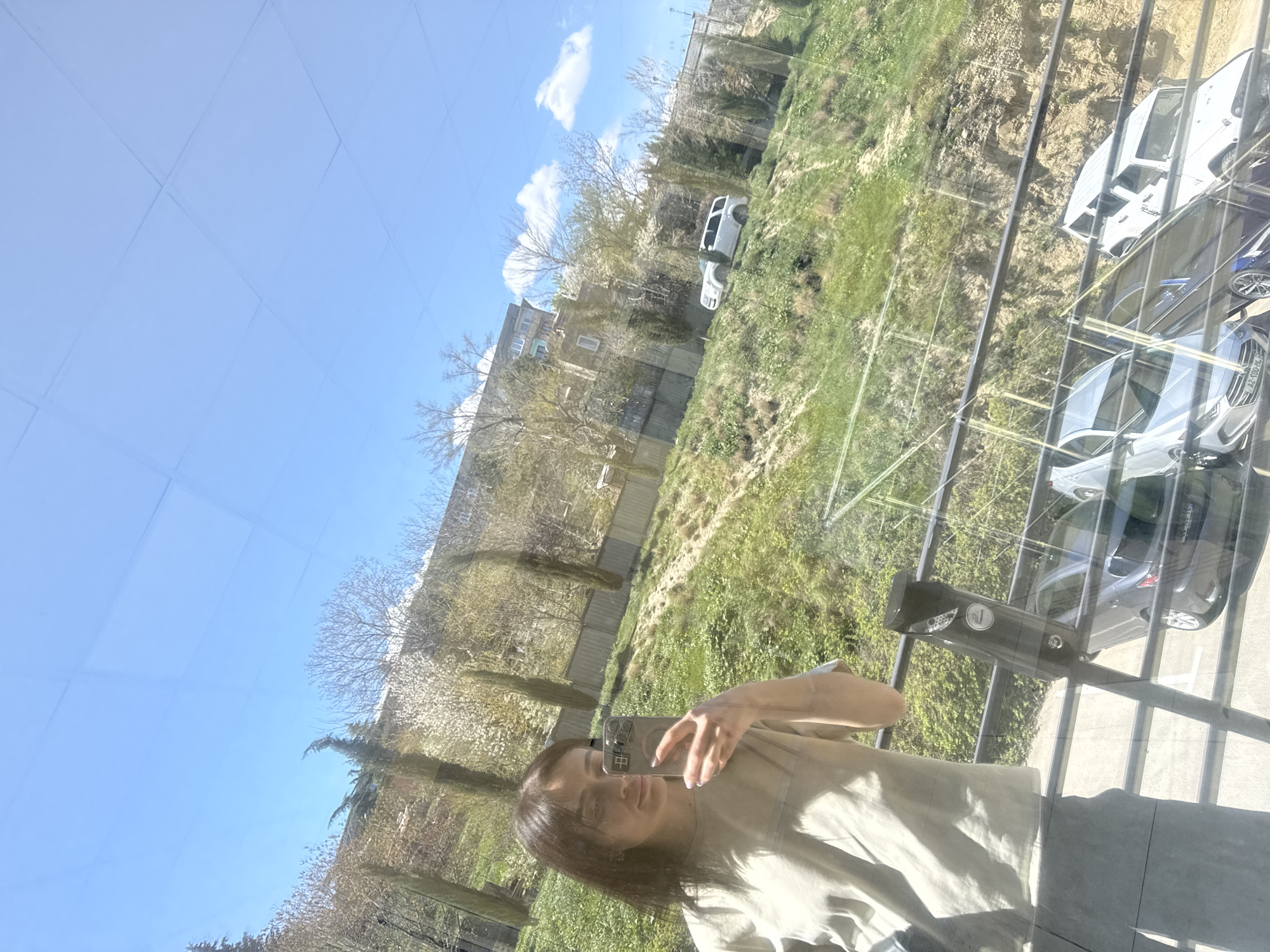 The image depicts a woman taking a selfie while inside a building. The view from the window shows a cityscape on a sunny day, likely in Tbilisi, Georgia, considering the architecture and landscape. The window reflection shows cars parked in a lot below, and there is greenery visible around the buildings and in the distance. The woman is holding her phone up to capture the image.