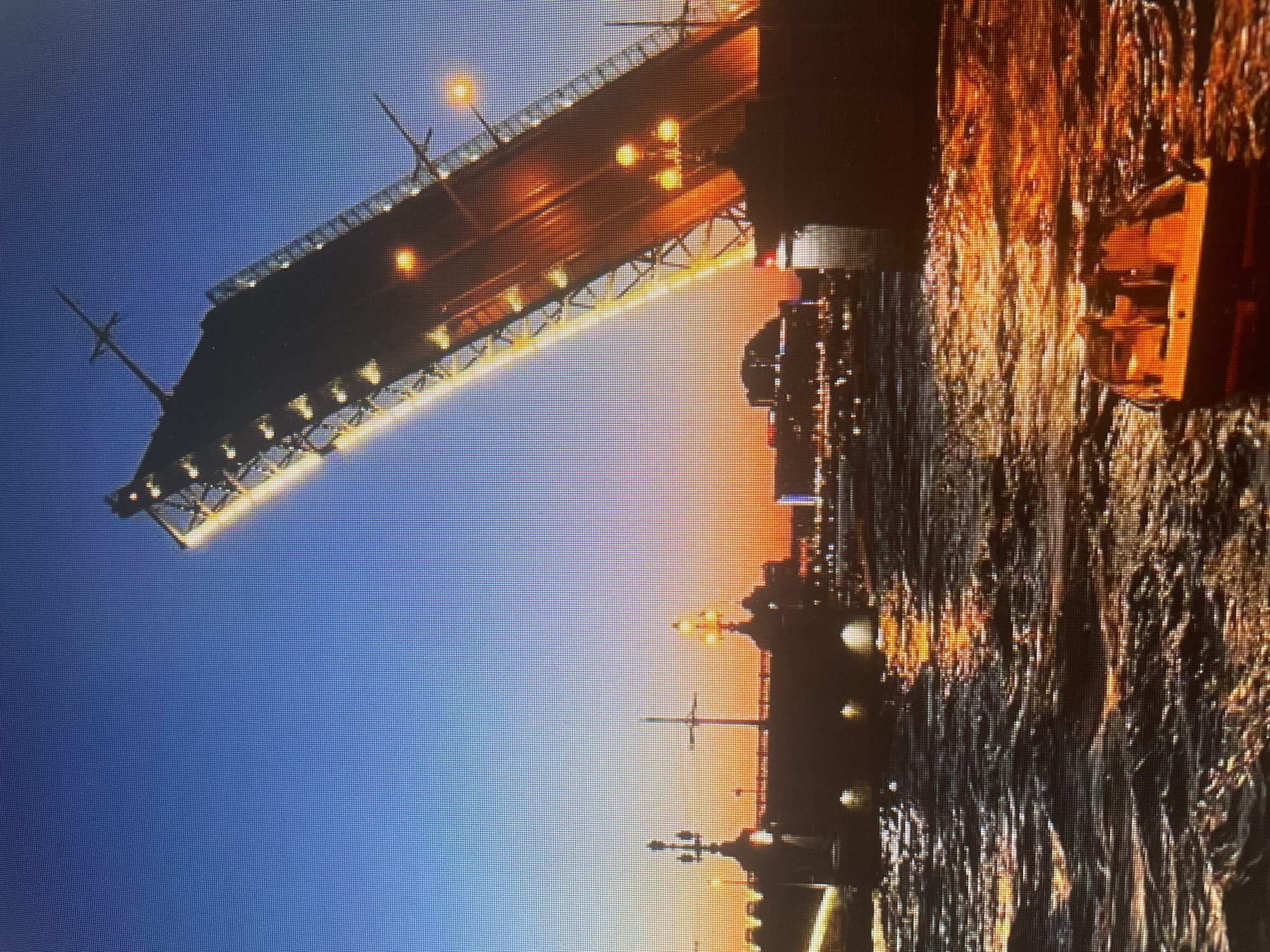 The image displays a view of a bascule bridge, likely the Sinyavino Bridge, in Russia. The bridge is elevated, suggesting it is open to allow water traffic to pass underneath. The sky transitions from a deep blue to a warm orange, indicating either dawn or dusk. The water below reflects the warm light of the sky and the bridge's lights, creating a shimmering effect. The bridge itself is illuminated with strings of lights along its underside and on lampposts, which are visible on the bridge and in the distance along the riverbanks. The cityscape in the background is silhouetted against the colorful sky, with more lights suggesting a developed urban or industrial area. The overall impression is one of a dynamic urban landscape at a transitional time of day.