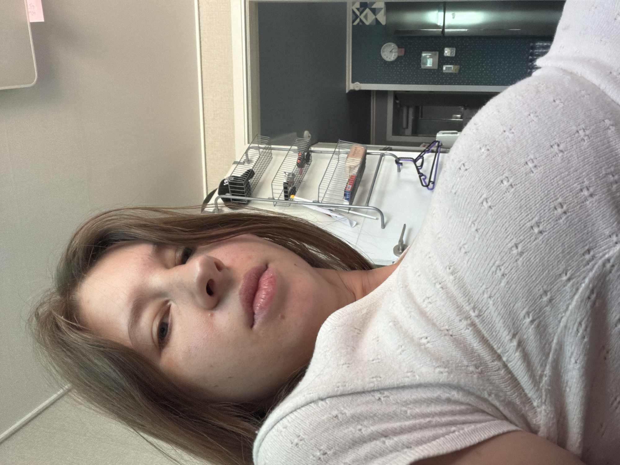 In a room with light-colored walls and beige baseboards, a young Caucasian female with light brown hair and fair skin is positioned in the foreground, looking towards the camera. She is wearing a light-colored, short-sleeved knit shirt with a textured pattern. Her expression is neutral, and her lips are slightly parted.
In the background, a doorway leads to another room. To the right of the doorway, a multi-tiered wire rack is attached to the wall, holding various items, including what appear to be makeup products and a baseball cap. Further in the background, on the right side of the image, a stainless steel refrigerator is visible against a dark teal wall with a subtle pattern. Above the refrigerator, a round clock with a pink accent is mounted on the wall. The overall setting suggests a domestic indoor environment, likely a home. The location is identified as Dongducheon-si, South Korea.