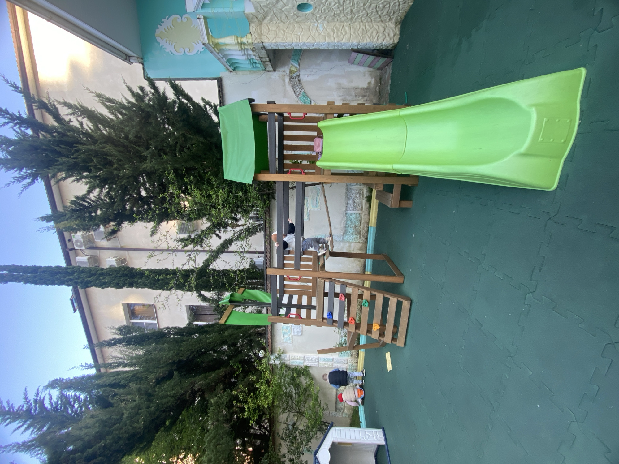 This image captures an outdoor play area in Sochi, Russia, bustling with children. The central feature is a wooden playset, stained dark brown, equipped with a bright lime-green plastic slide and a smaller green fabric canopy providing shade over a platform. The ground is uniformly covered with dark green interlocking foam mats, suggesting a safe play surface.
At least three children are visible, engaged in play. One child, wearing a light-colored top and gray pants, is positioned on the playset's wooden platform. Below, near the base of the playset, two other children are on the ground; one in a dark jacket and another wearing a light jacket and a red hat, seemingly occupied with objects on the mat. A small white and blue plastic playhouse is also present in the lower right corner.
The backdrop features several tall, dense evergreen trees, likely cypress, that frame the scene on the left and extend into the middle ground. Behind these trees, a light-colored building with multiple windows and external air conditioning units is visible. To the right of the playset, another building or wall features decorative light blue and yellow stucco with textured patterns. The soft, natural light indicates it is daytime, possibly late afternoon or early evening, under clear skies. There is no visible text in the image.