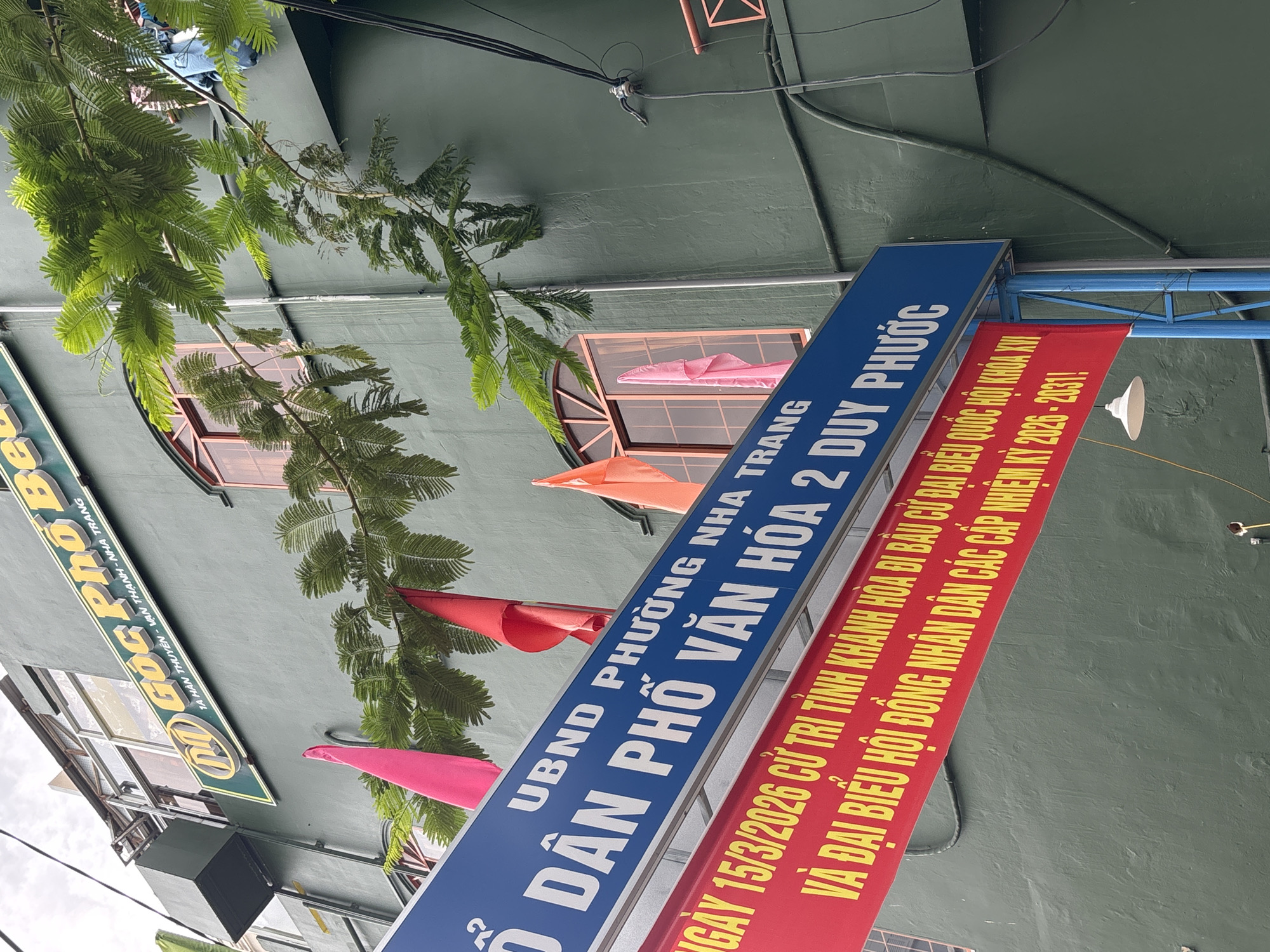 The image shows a green building with a large blue sign that reads "UBND PHUONG NHA TRANG DÂN PHÓ VĂN HÓA 2 DUY PHƯỚC". Below this sign, a red banner displays text in yellow, "NGÀY 15/3/2026 CỬ TRI TỈNH KHÁNH HÒA ĐI BẦU CỬ ĐẠI BIỂU QUỐC HỘI KHÓA XV VÀ ĐẠI BIỂU HỘI ĐỒNG NHÂN DÂN CÁC CẤP NHIỆM KỲ 2026 - 2031!". The building appears to be in Nha Trang, Vietnam. Several colorful flags are attached to the building, and a tree with green foliage hangs over the signs. The sky is overcast.