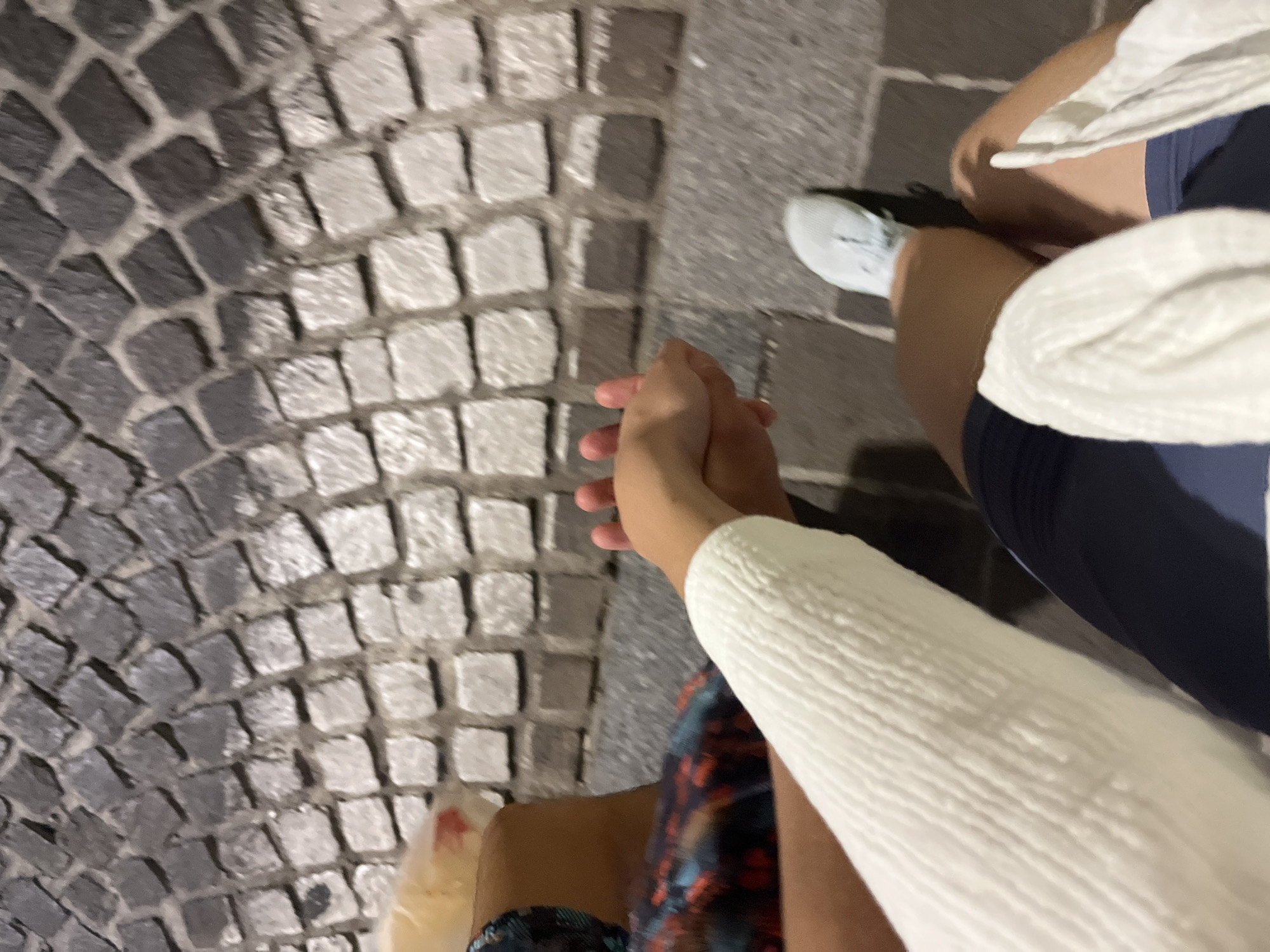 A slightly blurred, ground-level photograph shows two people holding hands while walking on a cobblestone street. The cobblestones are predominantly light grey and dark grey, forming a mosaic pattern. The person on the right wears white sneakers, dark blue shorts, and a white sweater. The person on the left wears sandals and patterned blue shorts. The hands are clasped in the center of the image. Based on the cobblestone pattern and general context, the location is likely in the Old Town of Budva, Montenegro.