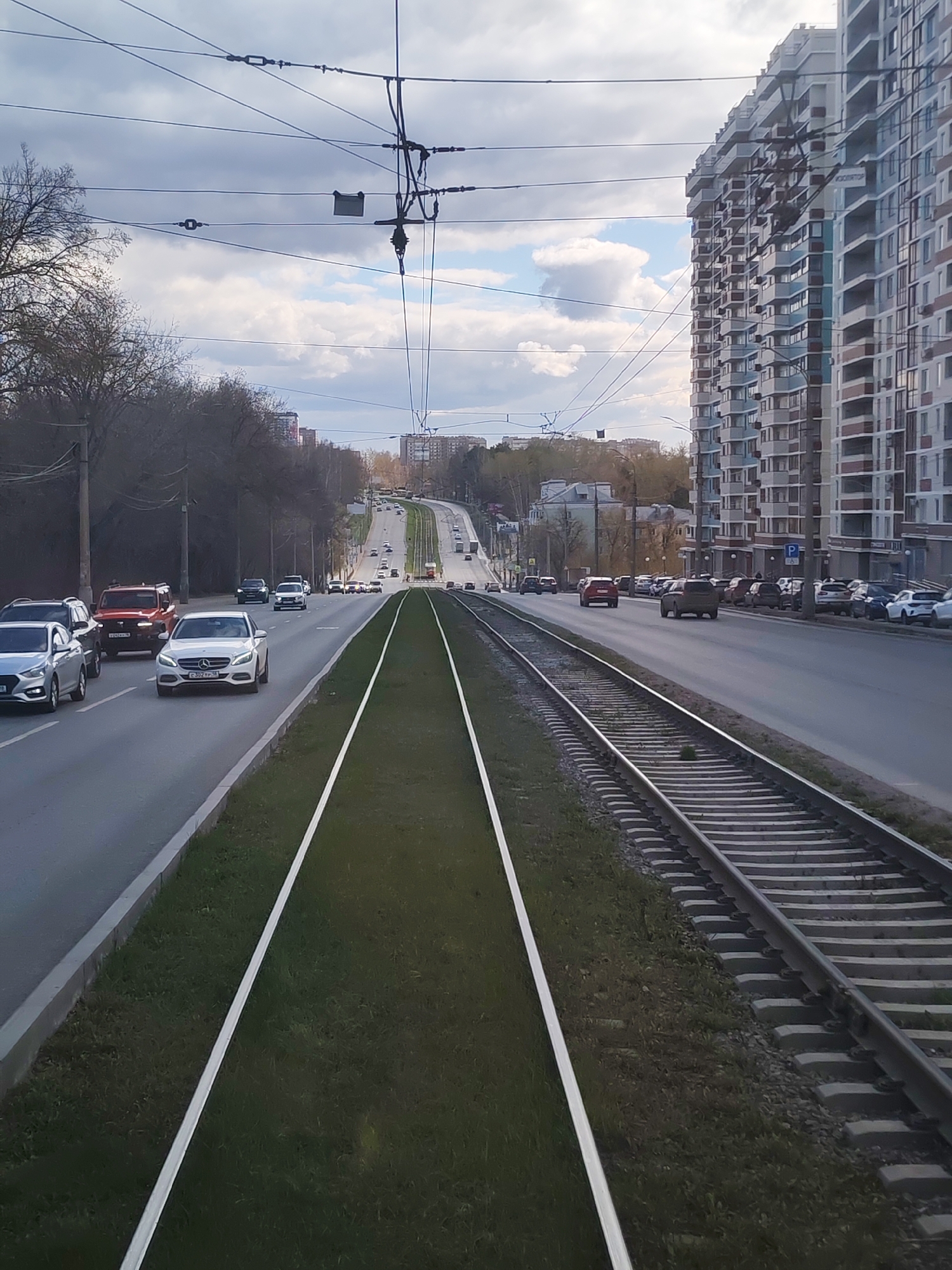 The image shows a street scene in Izhevsk, Russia, featuring a wide avenue with a tram line running down the center. The tram tracks are flanked by a strip of green vegetation and white lines. On either side of the tram line are lanes of vehicular traffic, with cars of various makes and models visible. The road ascends a slight incline in the distance. Buildings, including tall residential structures, line the sides of the avenue. Overhead, tram power lines and supports are visible against a backdrop of a cloudy sky. Trees are present along the sides of the road, indicating green spaces within the urban environment. The scene suggests a typical day with normal vehicular traffic.
