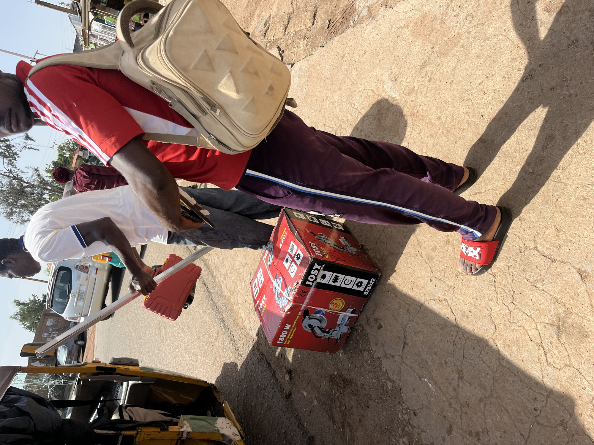 In Gura Top, Nigeria, two men are seen on a sun-drenched, cracked asphalt road. In the foreground, a person wearing a red shirt with white stripes, purple sweatpants with white and blue stripes, and red sandals, carries a tan shoulder bag. Next to them, a red box labeled "1800 W Heavy Duty Professional Miter Saw" is placed on the ground. To the left, a yellow auto-rickshaw, partially visible, has its rear door open, revealing bundled items and what appears to be a broken rear window. Another man, wearing a white t-shirt and blue jeans, is in the mid-ground, holding a red tool case and a long metal pole. In the background, a silver SUV and other vehicles are parked along the roadside, with buildings and trees lining the street.