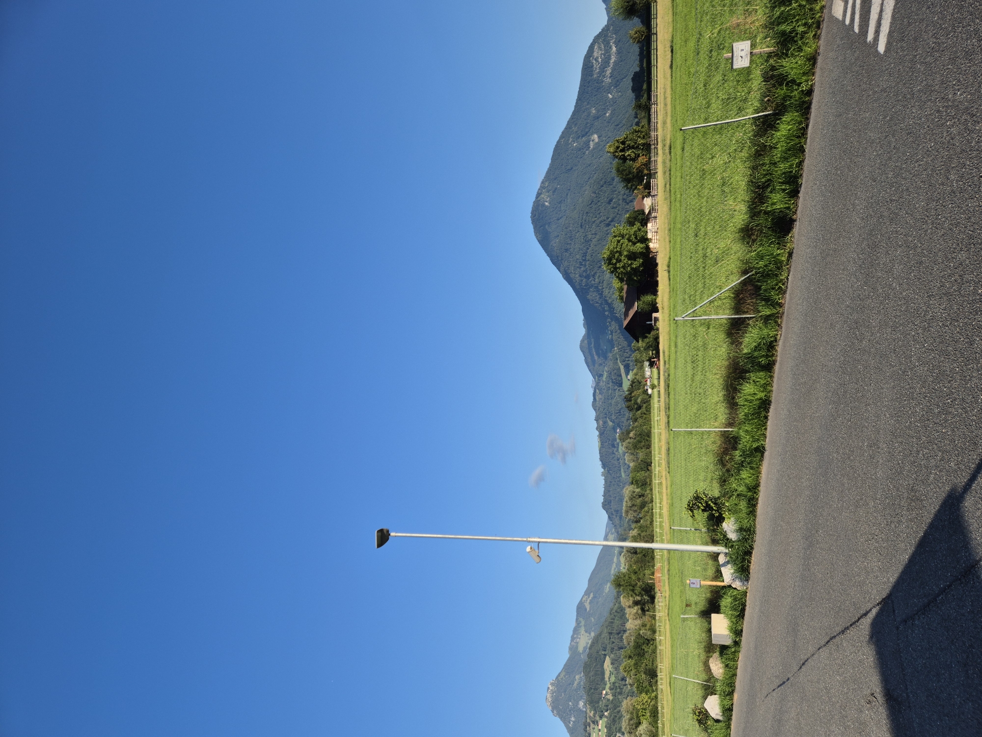 The photograph captures a sunlit outdoor scene near Niederndorf, Austria. The sky is a clear, vibrant blue, dominating the upper portion of the frame. In the mid-ground, a vast, green field stretches out, bordered by a fence. A tall, silver lamppost stands prominently in the field, with a smaller fixture attached to it. Behind the field, a cluster of trees forms a natural border, beyond which rolling hills and mountains rise, their slopes covered in dense green foliage. A few scattered buildings, including a dark-colored barn with a red roof, are visible nestled amongst the trees and on the hillsides. In the foreground, a grey asphalt road runs diagonally across the lower part of the image, with a distinct shadow cast across it from an unseen object. A white crosswalk marking is faintly visible on the road. Several small, informational signs are placed near the fence line in the field. The overall impression is of a tranquil rural landscape on a clear day.