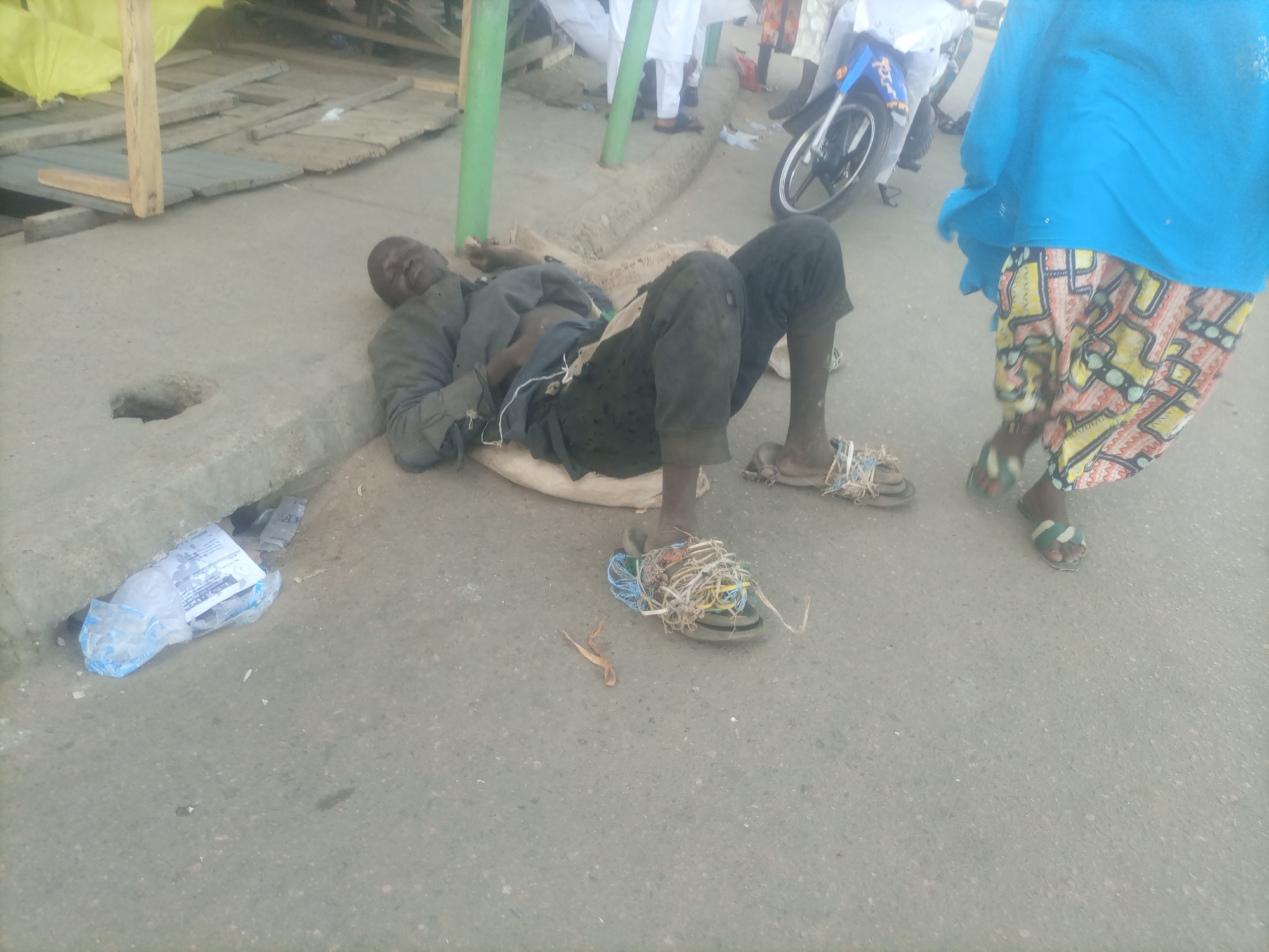 In Arawa, Nigeria, a man is lying on his back on a dirt and concrete surface next to a curb. He is wearing a dark long-sleeved shirt and dark pants. His feet are bare, except for one foot which has a sandal on it that is wrapped with various colorful ropes and wires. Next to him is a light-colored mat or blanket. In the background, several people are standing, some wearing traditional clothing. A blue motorcycle is parked behind the man. There is a yellow tarp stretched over a wooden structure in the upper left of the frame, and some scattered debris on the ground, including a plastic bag containing papers near the curb.