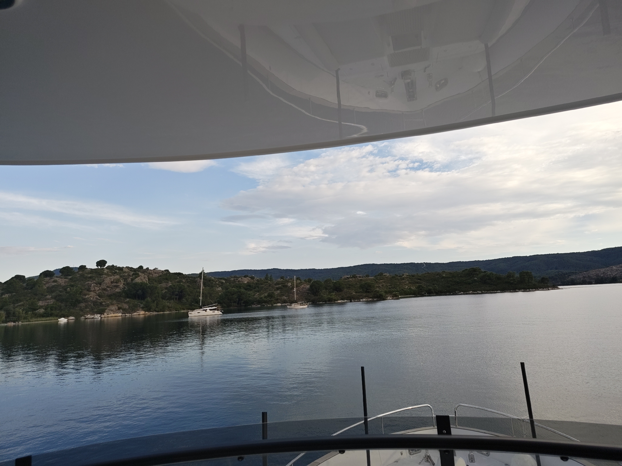 The image depicts a calm body of water, likely the sea, under a sky with scattered clouds. In the distance, a tree-covered, rocky coastline forms the horizon. Two sailboats are visible on the water, one closer to the shore and another further out. The perspective suggests the photo was taken from a boat, as the railing of a vessel is visible in the foreground. The overall scene is serene and suggests a location in a coastal area. Given the context of Sithonia Municipal Unit, Greece, this scene is characteristic of the coastal landscapes found in that region.