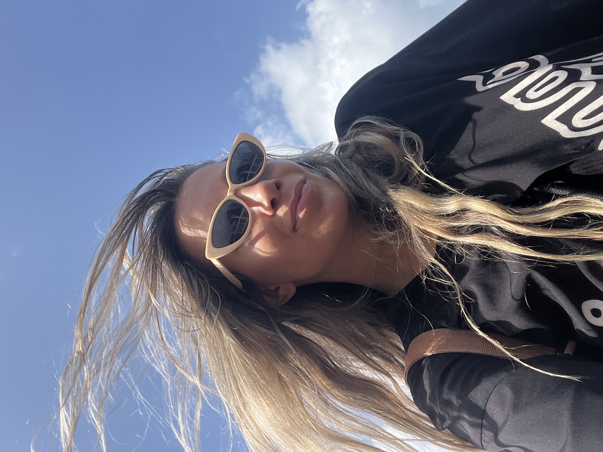 A low-angle, eye-level outdoor selfie features a young woman with long, blonde hair blowing in the wind. She is wearing cat-eye sunglasses with cream-colored frames and a black shirt with a graphic print. The background is a bright blue sky with scattered white clouds. The overall mood is casual and windswept, suggesting an outdoor activity in a coastal or open area, possibly in Walgama, Sri Lanka, given the clear blue sky and the presence of what appears to be a tropical climate. There are no other people or specific city landmarks visible.