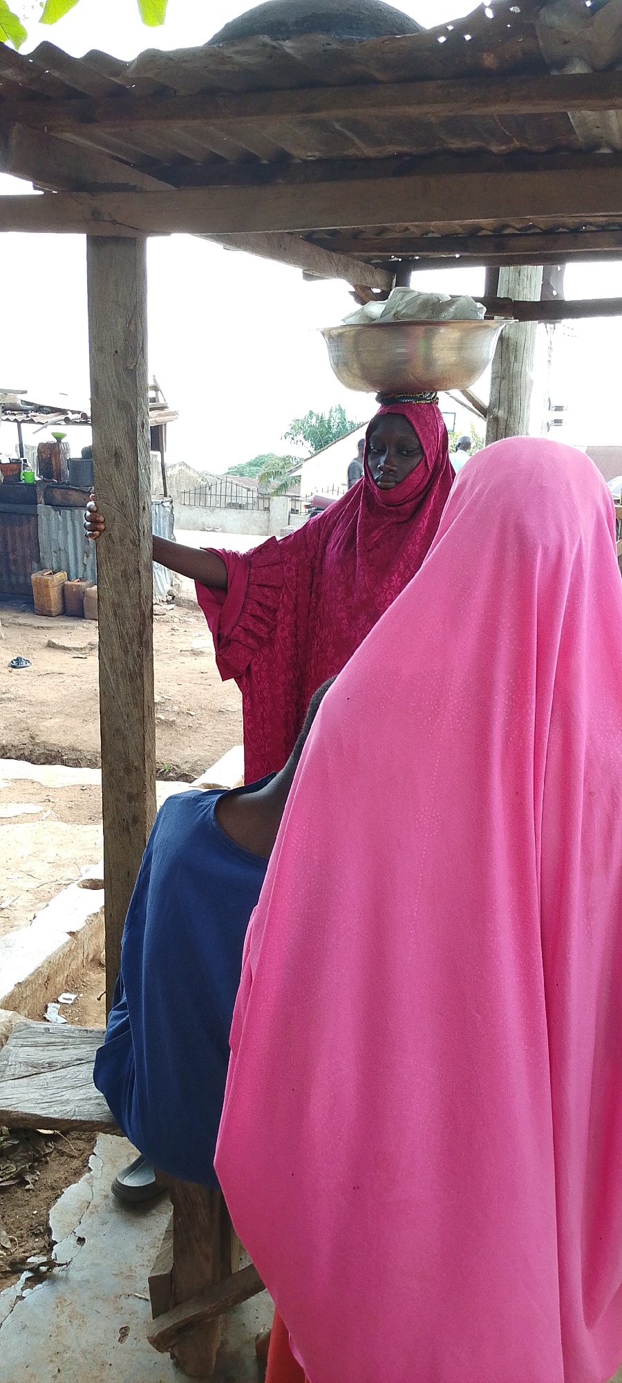 In Gumel, Nigeria, a woman wearing a maroon headscarf and matching flowing gown, with a large metal bowl balanced on her head, stands under a wooden shelter. She is leaning against a thick wooden support post, her left arm resting on it. Beside her, another person is obscured by a bright pink fabric, with only a blue long-sleeved shirt visible. The ground is a mixture of dirt and concrete. In the background, several stalls with corrugated metal roofs are visible, containing various containers and items, suggesting a market setting. The sky is overcast.