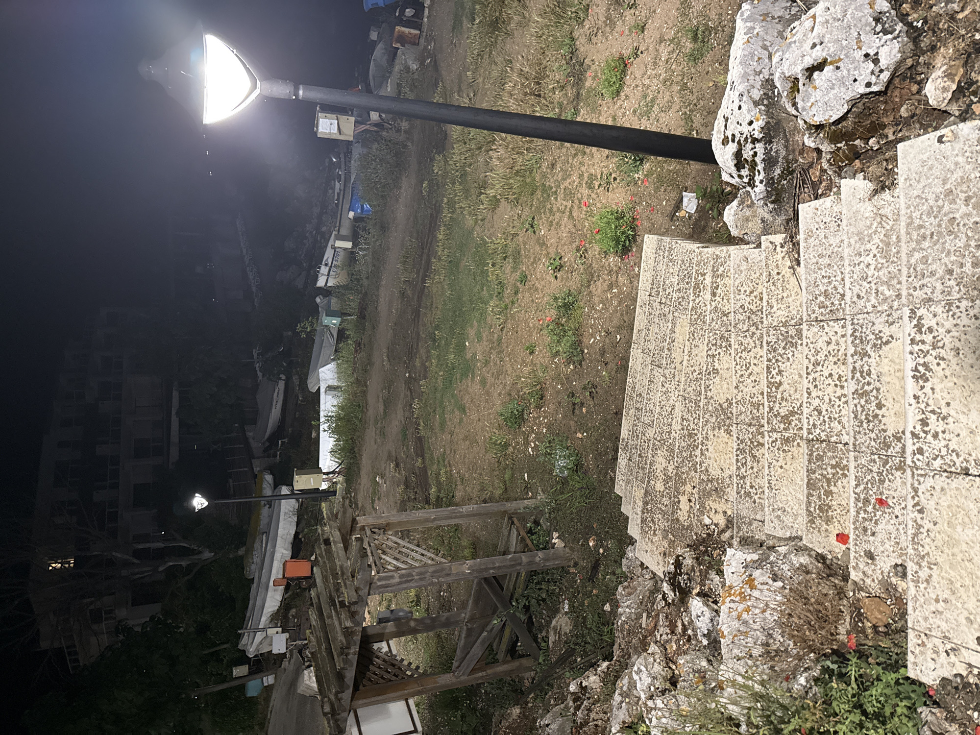The image depicts an outdoor scene at night in the city of Tyulenovo, Bulgaria. In the foreground, a set of stone stairs descends from the viewer's perspective. To the left of the stairs are large, moss-covered rocks and some greenery. Further into the scene, a weathered wooden gazebo or shelter is visible. Beyond the gazebo, the ground is dirt and patches of grass, with several boats resting on the shore. A tall, modern lamppost with a bright light illuminates the area. In the background, a dark, multi-story building is silhouetted against the night sky. The overall atmosphere is dark and somewhat desolate, suggesting a quiet, possibly coastal, area.