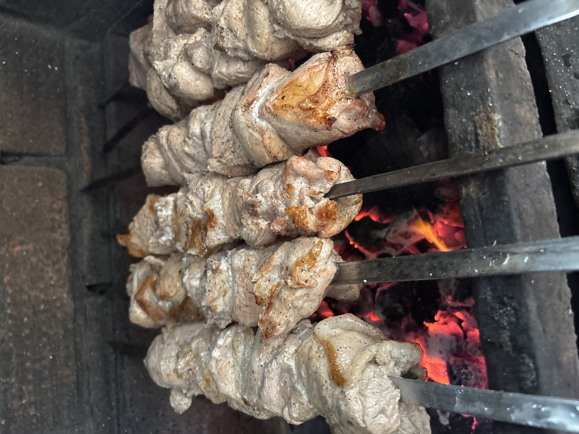 The image shows several metal skewers laden with pieces of meat, likely pork, cooking over a charcoal grill or fire pit. The meat is seasoned with black pepper or another dark spice, and the outer surfaces show signs of browning. The fire pit is constructed of dark, possibly brick or concrete materials. The image hints at an outdoor cooking or barbecue setting, but without specific context or details, the location in Chaikovsky, Russia, cannot be determined with certainty from this visual information alone. There are no people visible in the shot.