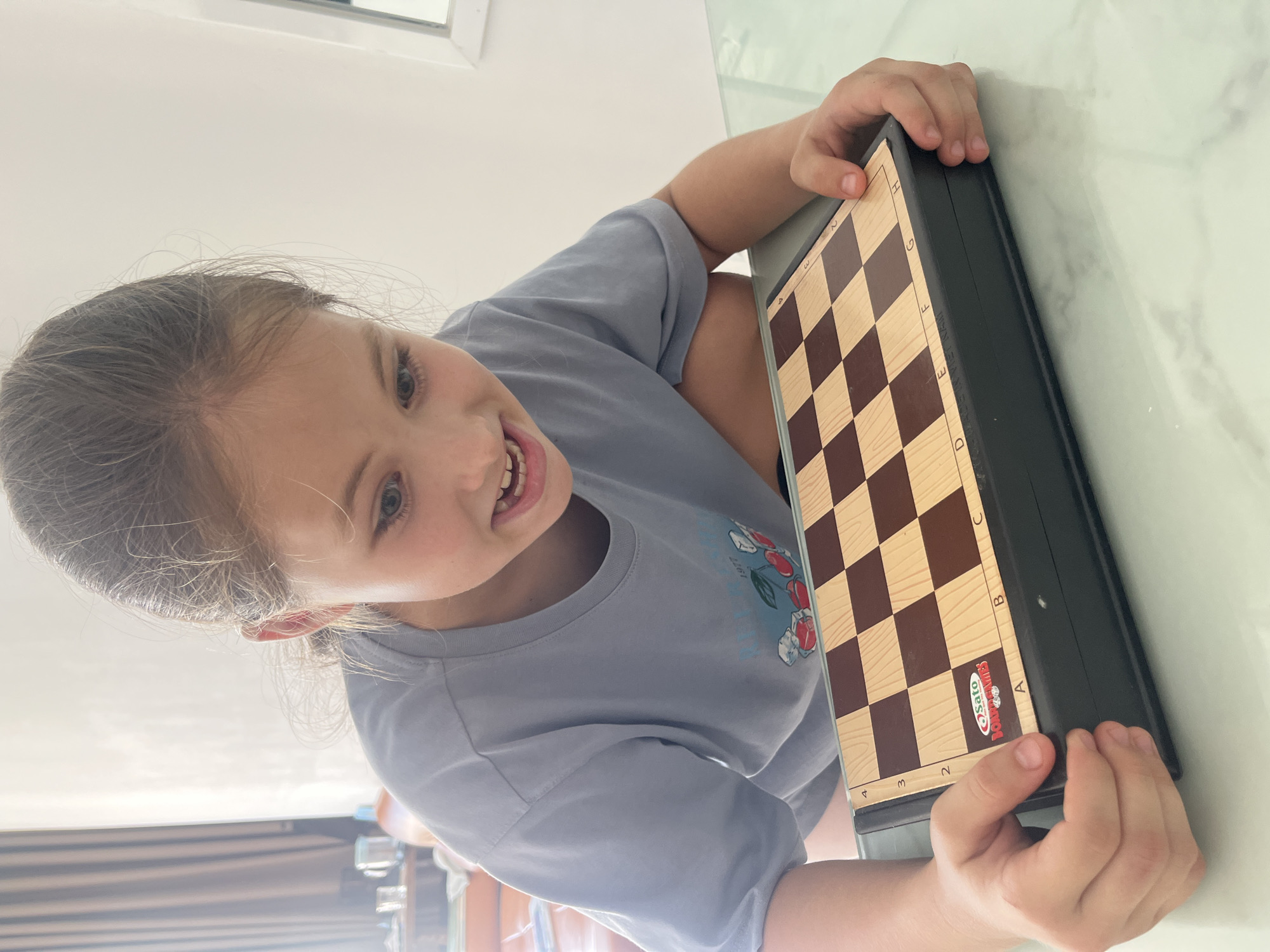 A young girl is holding a chessboard in front of her. The chessboard is brown and tan with black borders. The girl is wearing a blue t-shirt with a floral design. She is smiling and looking at the camera. The location is Bac Nha Trang Ward, Vietnam. The environment is a brightly lit interior space with white walls.
