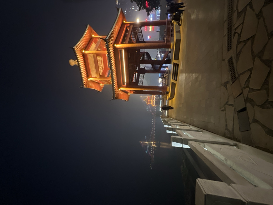 An evening scene in Jiangjin, China, depicts a traditional Chinese pavilion brightly illuminated on a riverside promenade. The two-tiered octagonal pavilion, constructed from reddish-brown wood with intricate details and a distinctive curved roof, dominates the right side of the frame, glowing with warm orange light. A spiral staircase is visible within its open lower level.
To the left, a light-colored stone balustrade lines a dark body of water, likely a river, which reflects distant lights. A lone individual, dressed in dark attire, walks away from the camera along the paved promenade between the balustrade and the pavilion. Further right, near the base of the pavilion, a small group of approximately three to four individuals is gathered, some appearing to sit on benches, with a small motorized scooter or similar vehicle parked nearby. City lights are faintly visible in the background beyond the pavilion on the right.
Across the water in the distance, a large suspension bridge is illuminated with a pattern of colorful lights, its reflection visible on the water's surface. Beyond the bridge, a smaller, similarly traditional pavilion-like structure is also discernible, illuminated. The sky is dark, indicating nighttime, and a slight atmospheric haze or fog is present, particularly over the water and in the distance. The promenade surface consists of irregularly shaped flagstones transitioning to smoother concrete sections. Drainage grates are present in the foreground paving.