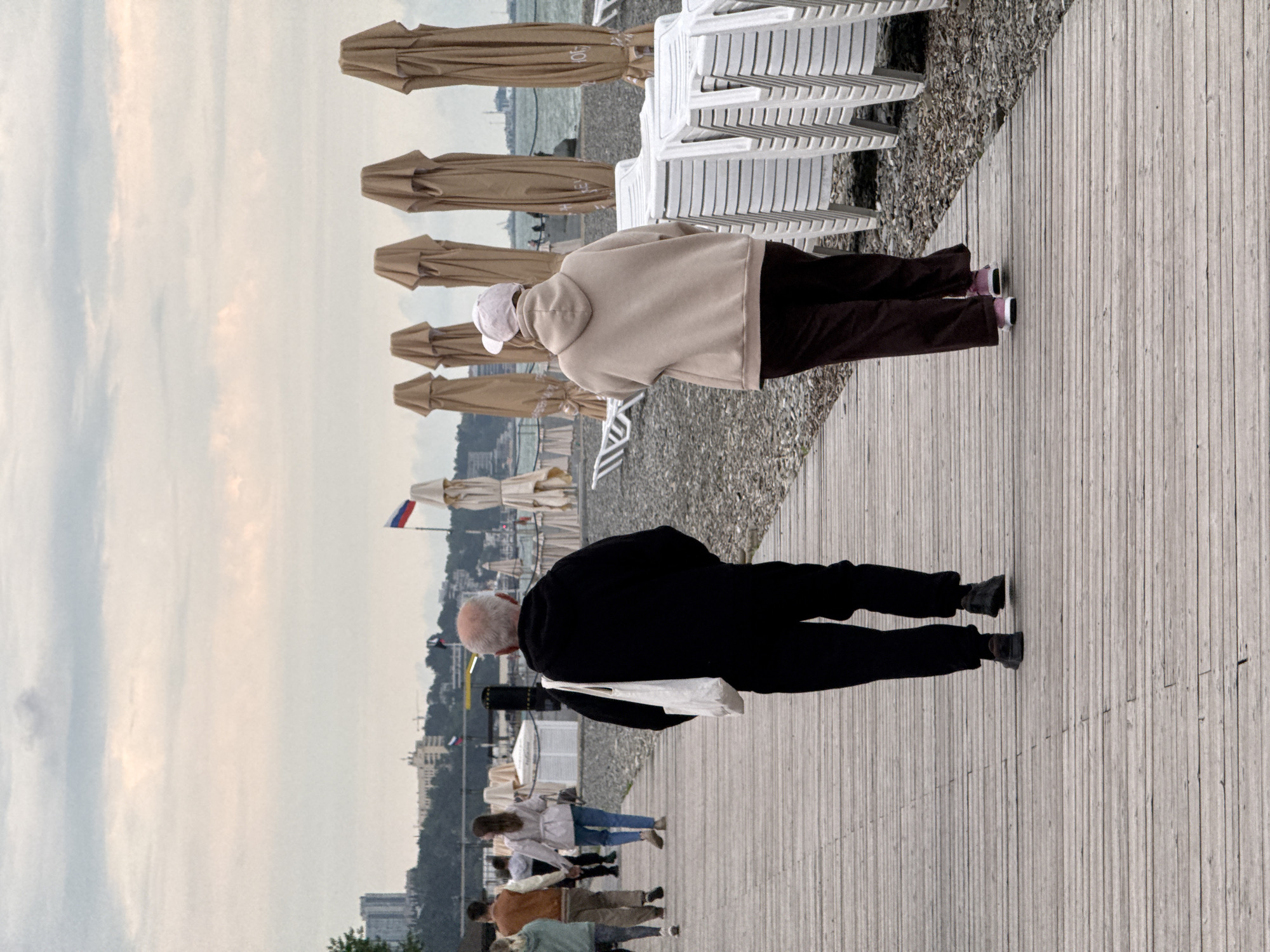 The image captures a scene at a beach in Sochi, Russia. A boardwalk stretches along the foreground, with several people walking on it. An elderly man in a black jacket is taking a photo towards the city. A woman in a beige hoodie and dark pants walks toward the beach. Folded umbrellas and stacks of white chairs line the beach beside the boardwalk. In the distance, buildings are visible and a Russian flag waves. The sky is overcast.