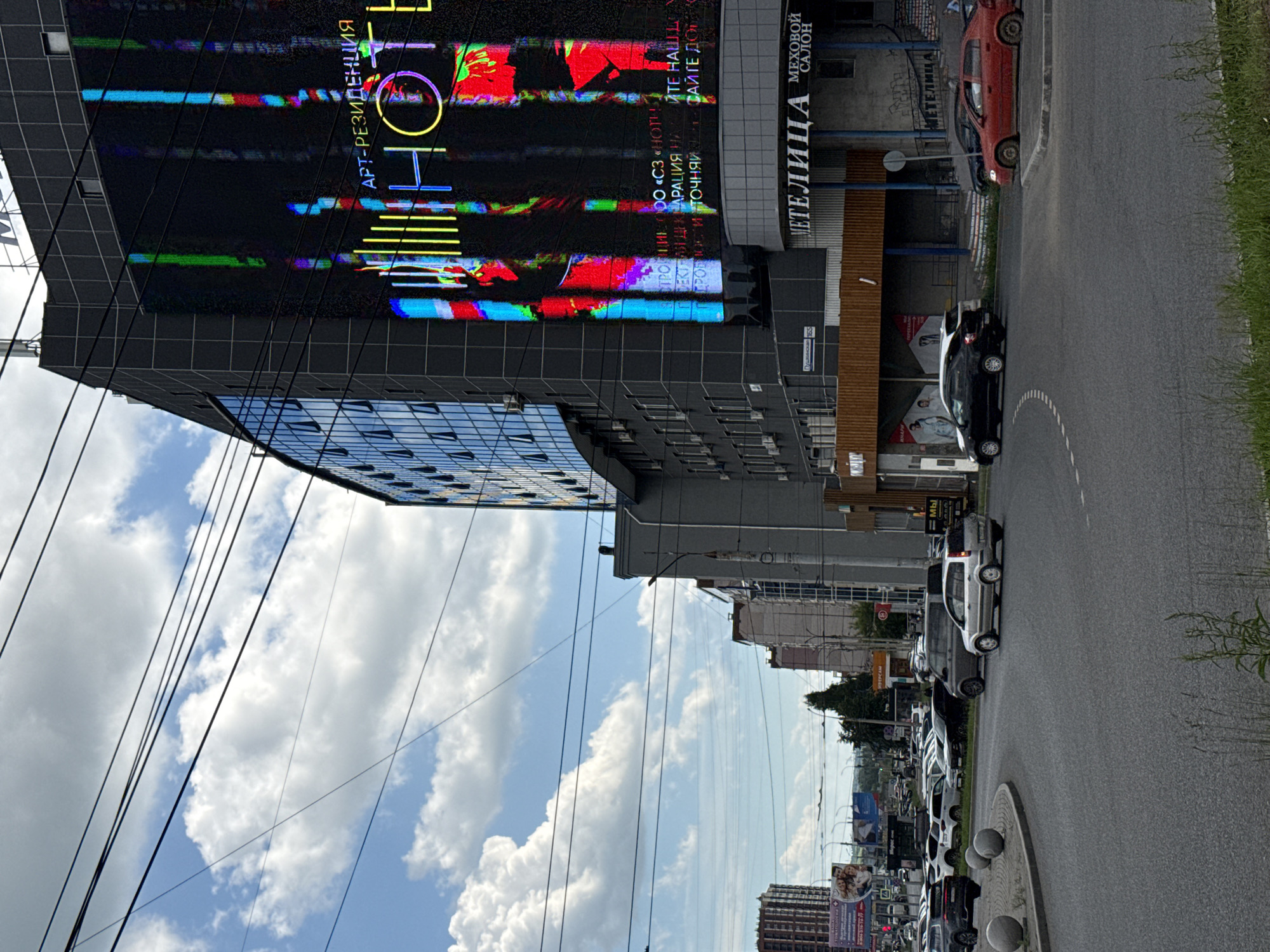 This is a street-level, vertical photograph taken in Izhevsk, Russia, likely during the daytime, given the bright sky and clouds. The scene captures a modern building with a large, illuminated digital billboard displaying colorful, abstract patterns and text. The text, predominantly in Russian, includes "АРТ-РЕЗИДЕНЦИЯ" (ART-RESIDENCY), "ООО «СЗ «НОТИВ»" (LLC "SZ" NOTIV"), and "МЕТЕЛИЦА МЕХОВОЙ САЛОН" (METELITSA FUR SALON). The building itself appears to be a commercial or residential complex, characterized by dark, grid-like panels and reflective glass windows.
The street in the foreground is paved and curves to the right. Several cars are parked along the side of the road and driving on it. A red car and a dark sedan are prominent. The sidewalk area features a circular planter with decorative spheres. The overall setting suggests an urban environment with a mix of modern architecture and everyday city life. The sky is partly cloudy, with fluffy white clouds against a blue background, indicating fair weather. The image's orientation is rotated 90 degrees counter-clockwise, making the sky appear on the left and the street on the bottom.