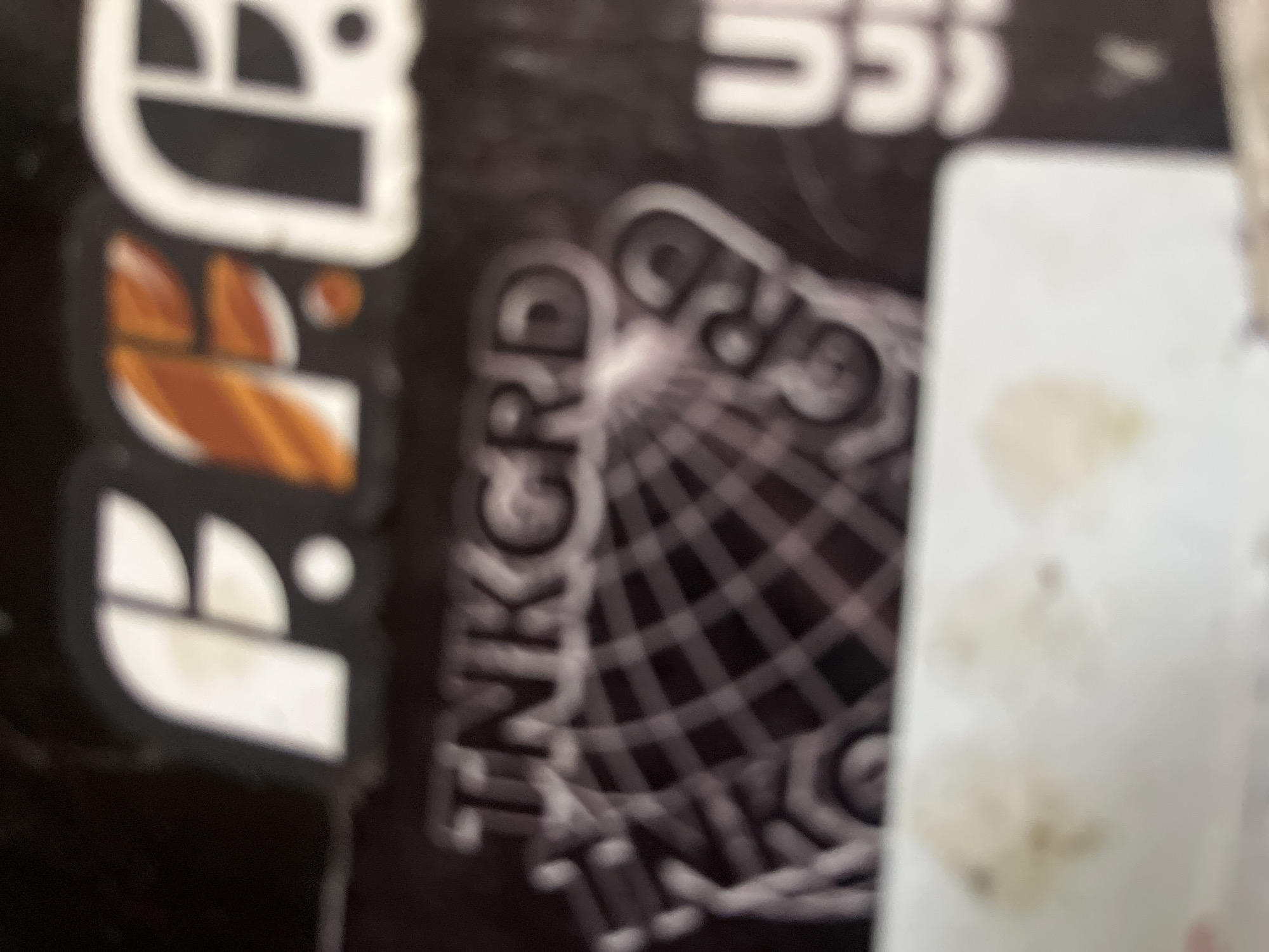 The image shows a close-up of a sticker or label with text and graphics. At the top, there are three stylized letters that appear to be "FFF" in white and orange. Below this, the text "TNKGRD" is visible in a distressed, stylized font, superimposed over a graphic resembling a net or globe. To the right, the letter "U" is partially visible, followed by what appear to be two "C"s. The background is dark. There are also smudged, lighter colored areas at the bottom of the image. The scene is indoors with no discernible environmental features. There are no people or actions depicted.