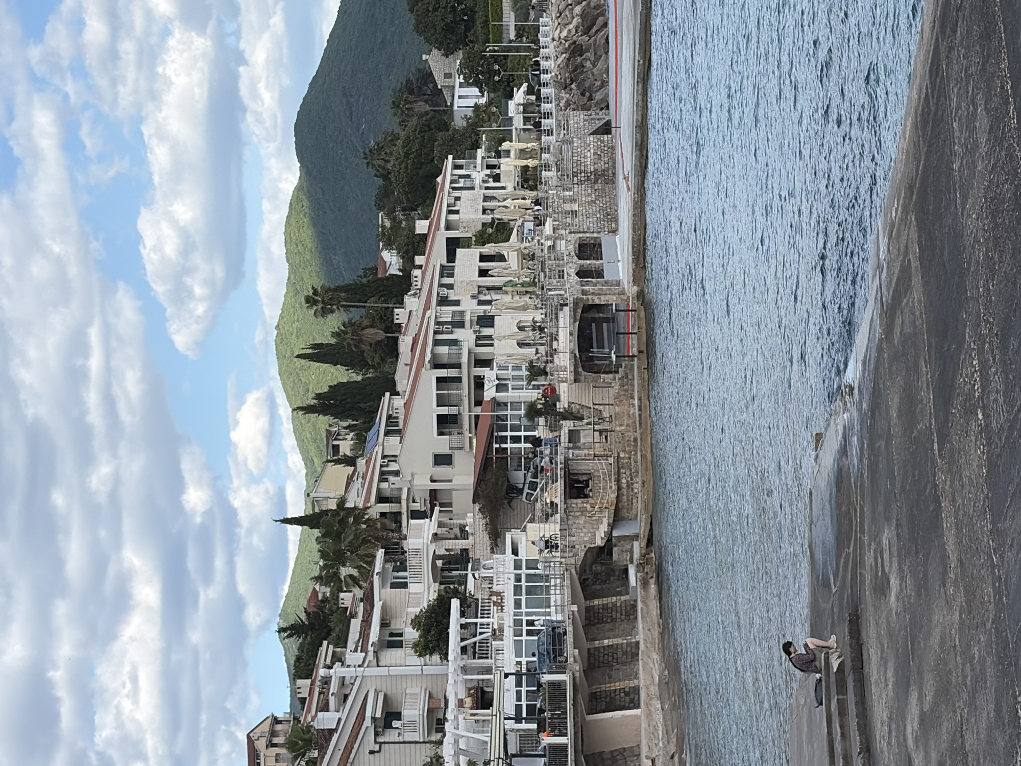 The image depicts a coastal view, likely in Herceg Novi, Montenegro, as evidenced by the architecture and landscape. Several multi-story buildings with white facades and balconies are situated along the coastline. Lush, green mountains rise in the background, partly obscured by scattered clouds in a bright blue sky. In the foreground, a stone pier or walkway extends into the water. A person is sitting on the edge of the pier, near the water's edge. The sea is calm and blue.