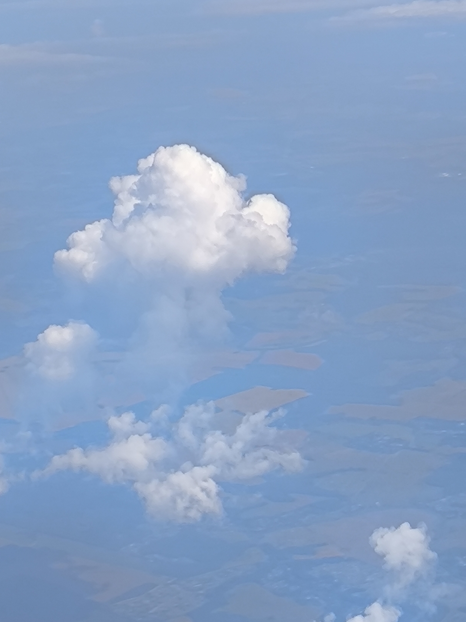 An aerial photograph captures a sky filled with scattered white cumulus clouds against a pale blue backdrop. Below the clouds, a landscape is visible, appearing as patches of muted colors suggestive of fields or land formations, possibly indicating a rural or semi-rural area. There are no discernible city elements or any signs of human activity in the visible portion of the scene. The overall impression is one of a clear day from a high vantage point, with a focus on the atmospheric conditions and the earth's surface from above.