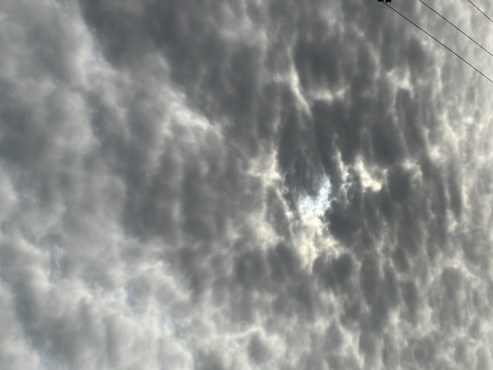 The image shows a sky filled with dark, heavy clouds, with a bright patch of sunlight breaking through in the center. The sun's rays create a luminous halo effect as they pierce the cloud cover. The clouds are textured and appear dense, suggesting a potentially overcast or stormy condition. In the lower right corner of the image, two dark electrical wires are visible, stretching diagonally upwards. These wires suggest the presence of man-made infrastructure in the environment. The overall impression is one of a dramatic sky, possibly indicating a shift in weather. The scene is likely an outdoor environment in Igboefon, Nigeria, given the context. There are no people or discernible actions taking place.