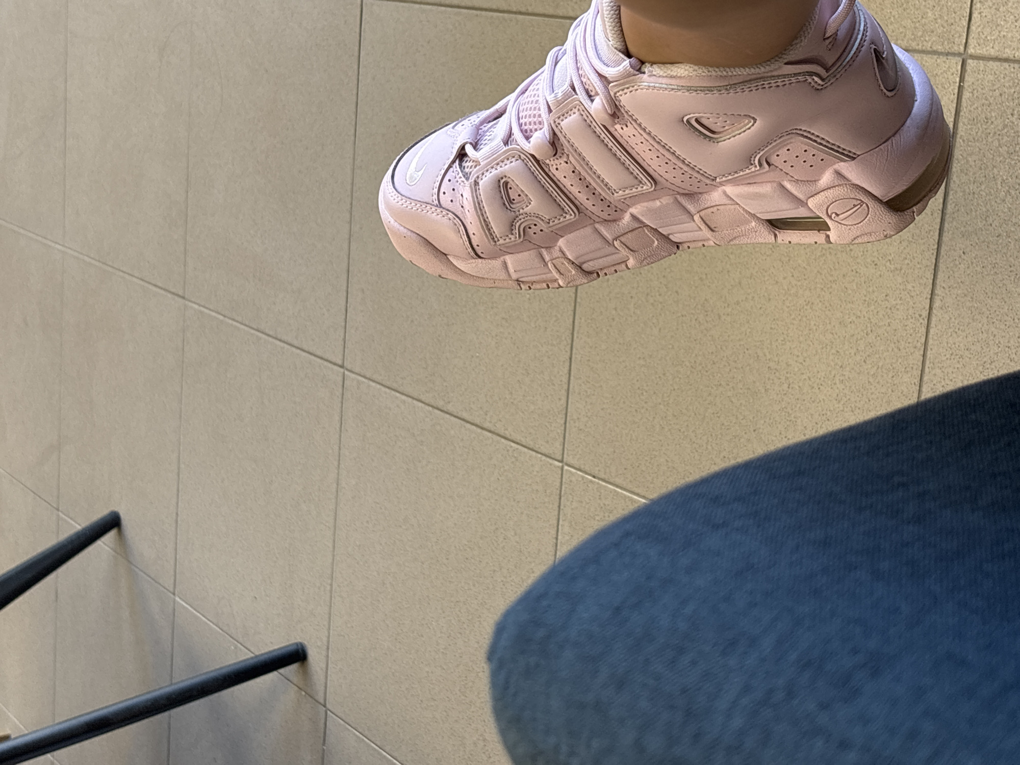 The image displays a close-up view of a person's legs and feet, placed on a light beige tiled floor. The person is wearing blue denim jeans and a light pink Nike sneaker with visible branding. The sneaker features a chunky design with multiple overlays and perforations. In the upper left portion of the image, two dark, thin legs of a chair or table are visible, resting on the tiles. The scene appears to be indoors, possibly in a public space like a waiting area or a café. The lighting suggests it is daytime. There is no specific indicator of the location being in Plovdiv, Bulgaria.