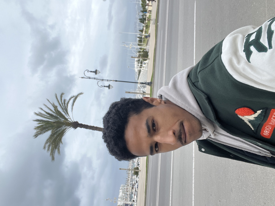 A young adult male is depicted in a medium close-up, facing slightly towards the viewer's right and looking directly at the camera. He has dark, curly hair and a light mustache, and is wearing a light grey hooded sweatshirt beneath a dark green and white varsity-style jacket. The jacket features an embroidered patch on the left chest depicting a stylized mountain with a red sun, accompanied by the text "ORIGIN RANGER," and partial green lettering on the right sleeve.
The scene is set in Tanger, Morocco, specifically along a waterfront promenade adjacent to a marina. Behind the subject, a paved multi-lane road is visible, beyond which numerous white boats and yachts are docked in the marina. Several white buildings line the waterfront. A tall palm tree with green fronds stands in the mid-ground, positioned to the left of an ornate black street lamp with two lights. The sky is uniformly overcast with grey cloud cover, and distant landmasses are faintly visible on the horizon.