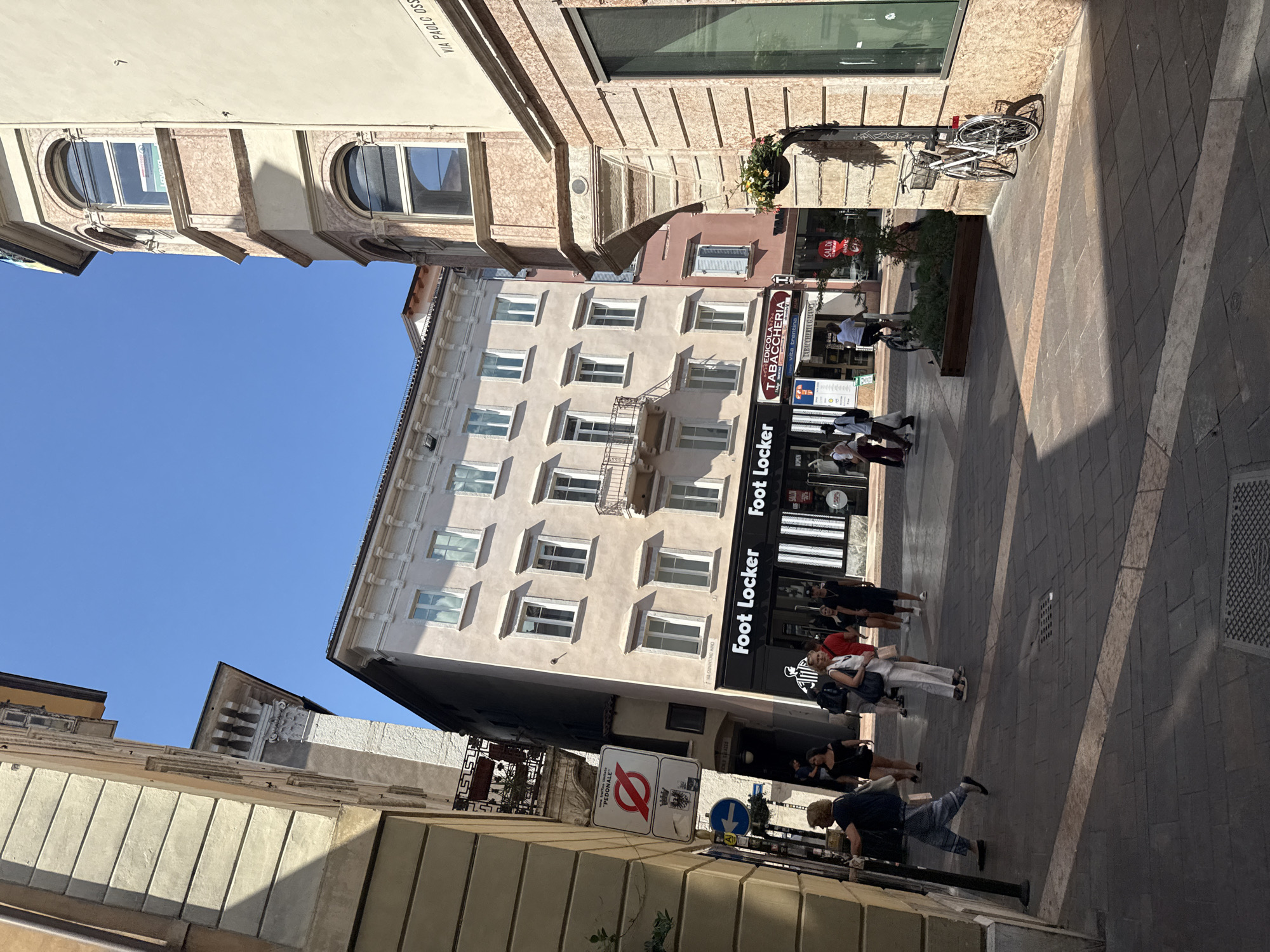 The image captures a street scene in the city of Trento, Italy. The perspective is looking down a pedestrianized street lined with buildings. On the left, a tan building with distinct horizontal architectural lines is prominent. Further down the street, a tall, slender building with light-colored facades and multiple windows is visible, leaning slightly. A black awning with white text reading "Foot Locker" spans across the front of one of the buildings. Adjacent to Foot Locker, a "TABACCHERIA" sign is displayed.
Several people are walking along the street. Some are individuals, while others appear to be in small groups. A person is walking on the left side of the street, facing away from the camera. Further down, a group of people are walking towards the camera, with one individual in the foreground wearing a white top and light-colored pants.
A white bicycle is parked on the right side of the street, leaning against a building with a textured facade. The street is paved with dark cobblestones. Shadows cast by the buildings and people create strong contrasts across the pavement. A street sign with a red circle and a diagonal line across a symbol is visible on the left side of the street, indicating a pedestrian zone. Another blue sign with a white arrow points to the left. The sky above is clear and blue.