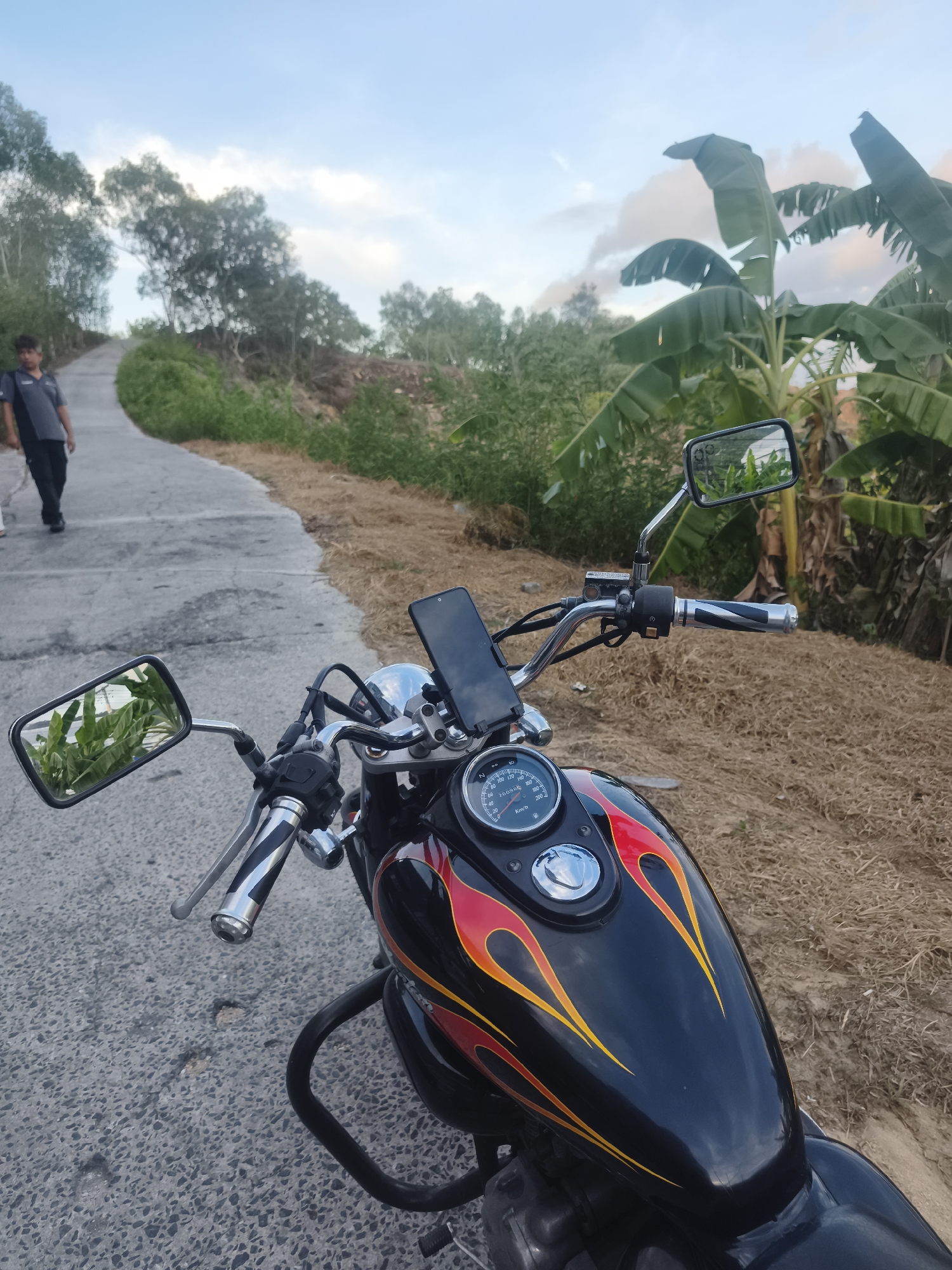 A man walks up a paved road in a rural area of Ko Samui District, Thailand, with a motorcycle parked in the foreground. The motorcycle is black with flame decals and has a phone mounted on the handlebars. Banana trees and other vegetation line the road. The sky is cloudy and appears to be late afternoon or early evening.