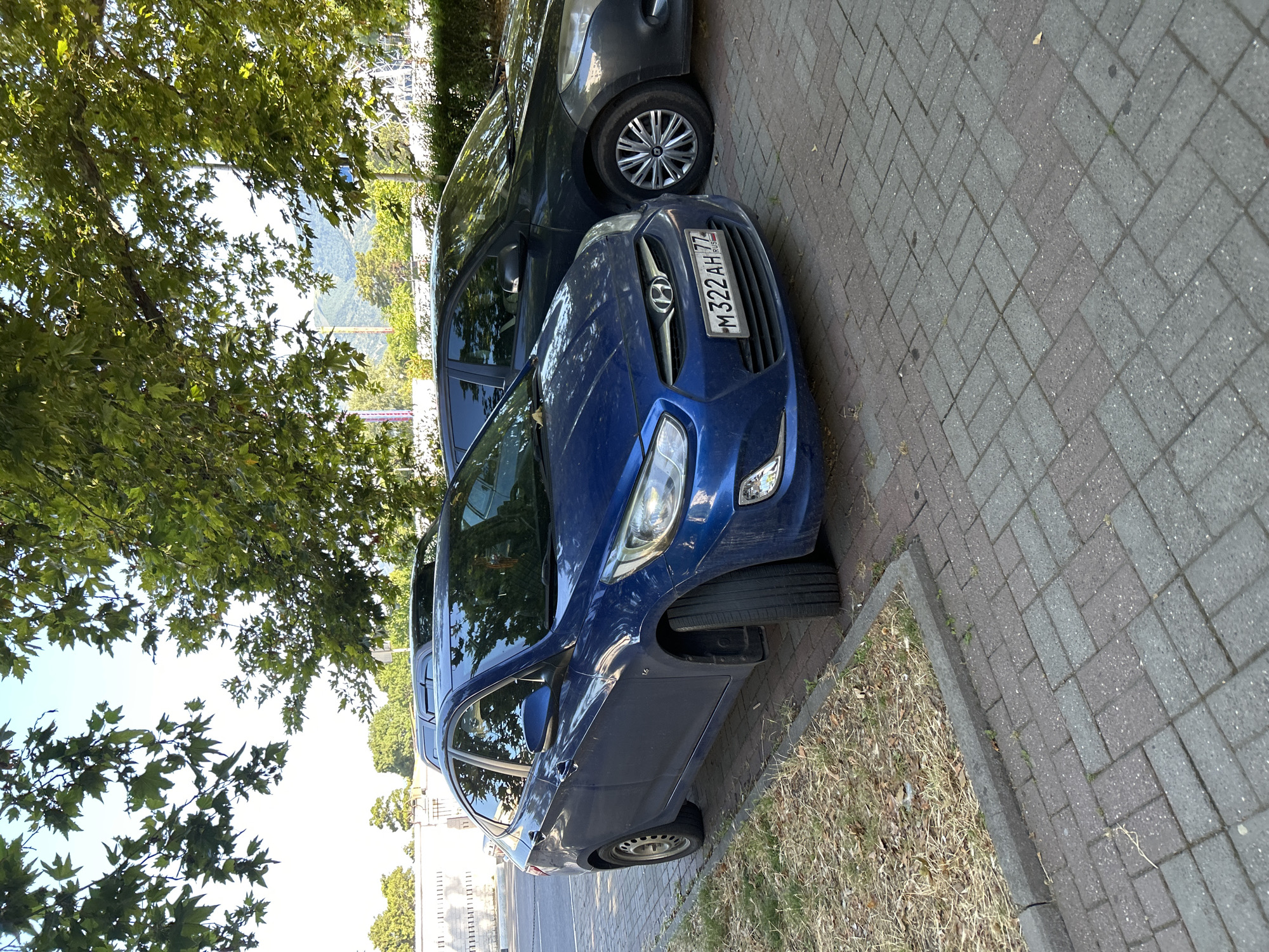 A blue Hyundai Solaris sedan is parked on a brick-paved surface next to a dark grey sedan. The blue car is in the foreground, facing slightly to the right. Its license plate, M322AH77, is visible. The car is positioned on the right side of the frame, with its front bumper and headlights in clear view. A patch of dry grass and a curb are visible to the left of the car. The grey sedan is parked to the right of the blue car, partially obscuring it. Trees with green leaves overhang the scene from above, casting shadows. In the background, a white fence and buildings are visible, along with distant hills. The setting appears to be a street or parking area in Gelendzhik, Russia, under a bright sky.
