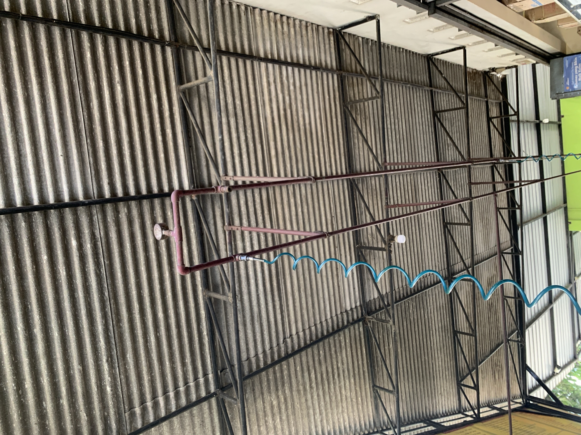 This is an outdoor shot of a corrugated metal roof structure supported by a dark metal frame. A blue coiled hose hangs from a red pipe system that is also attached to the roof structure. A pressure gauge is visible on the red pipe. The scene is likely a covered area, possibly a car wash or workshop, given the presence of the hose. The location is identified as Karangkemiri, Indonesia.