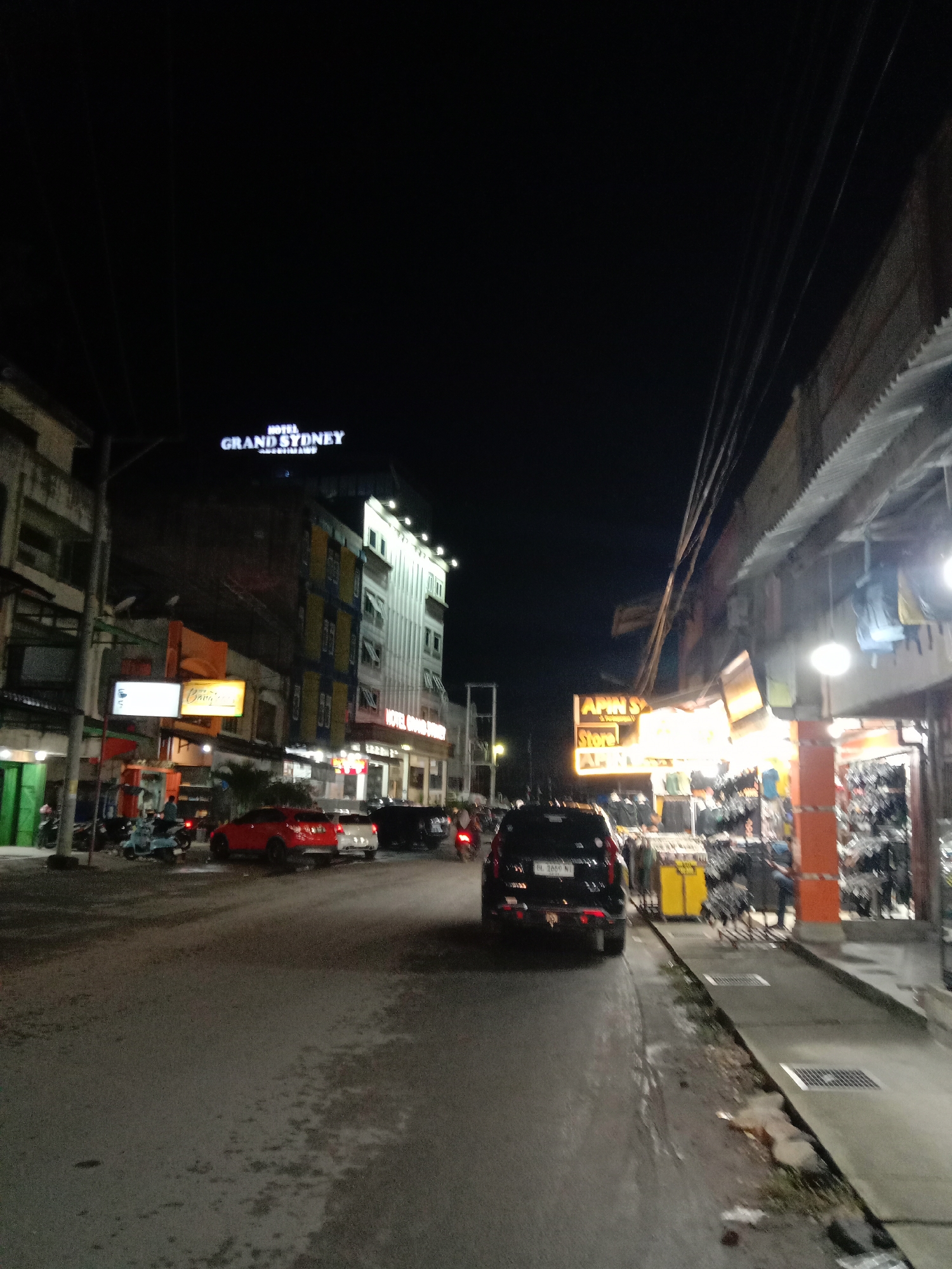 The image depicts a street scene at night in Lhokseumawe, Indonesia. "HOTEL GRAND SYDNEY" is illuminated atop a multistory building in the distance. A black SUV is visible in the middle of the street, facing away from the camera. Several parked cars are present, as well as motorcycles. On the right side of the image, there is a shopfront lit with yellow lights and selling clothes. Streetlights illuminate the area. Overhead power lines are visible. The road surface appears wet.