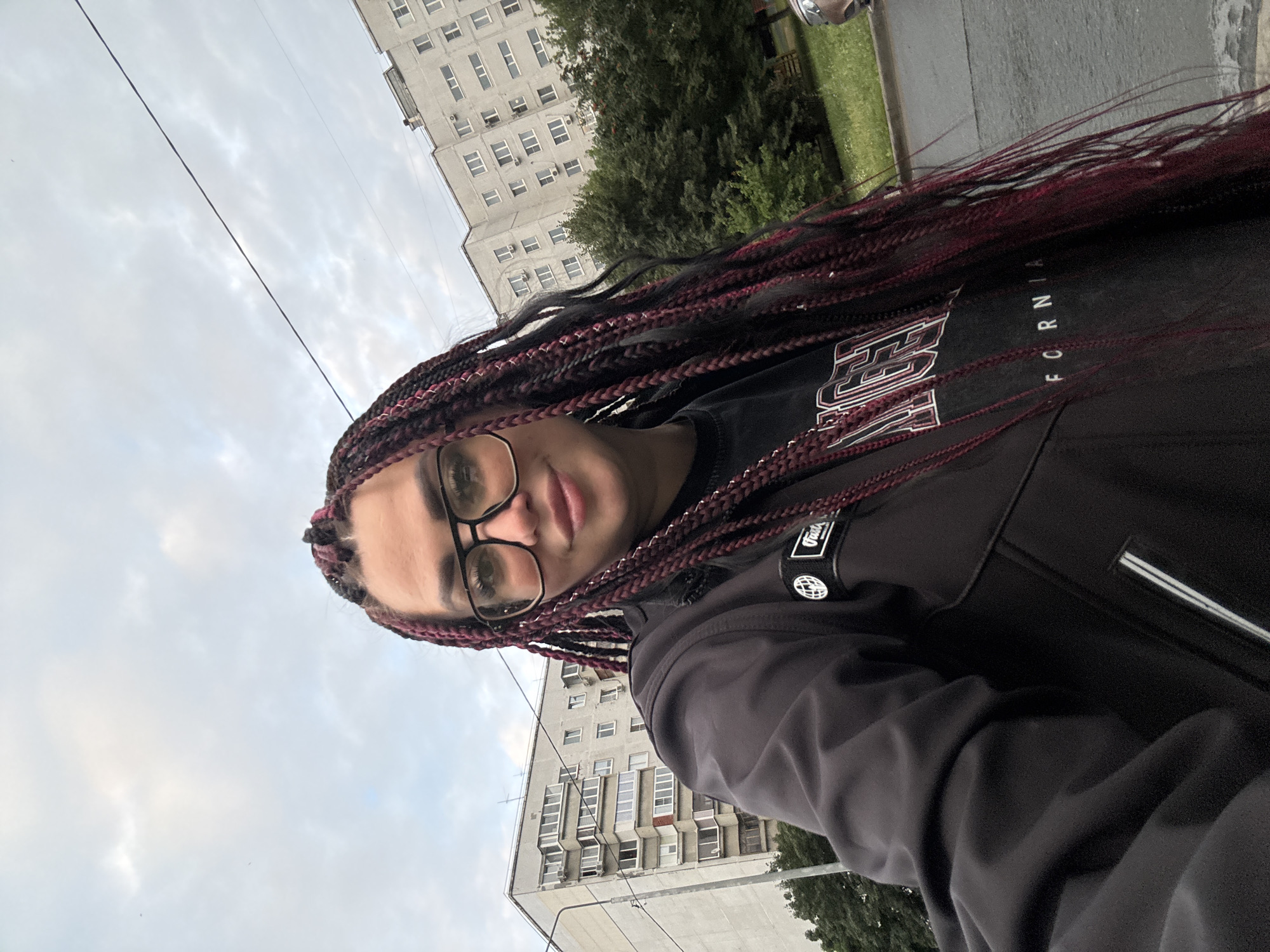 A selfie is taken from a low angle in an outdoor urban setting. The subject, a woman with braided hair, wears a black jacket over a t-shirt with red and white lettering. She has on large-rimmed glasses and is smiling at the camera. In the background, there are several multi-story apartment buildings, trees, and a street. The sky is overcast. The overall scene suggests a residential area in Tolyatti, Russia, likely taken on a cloudy day.