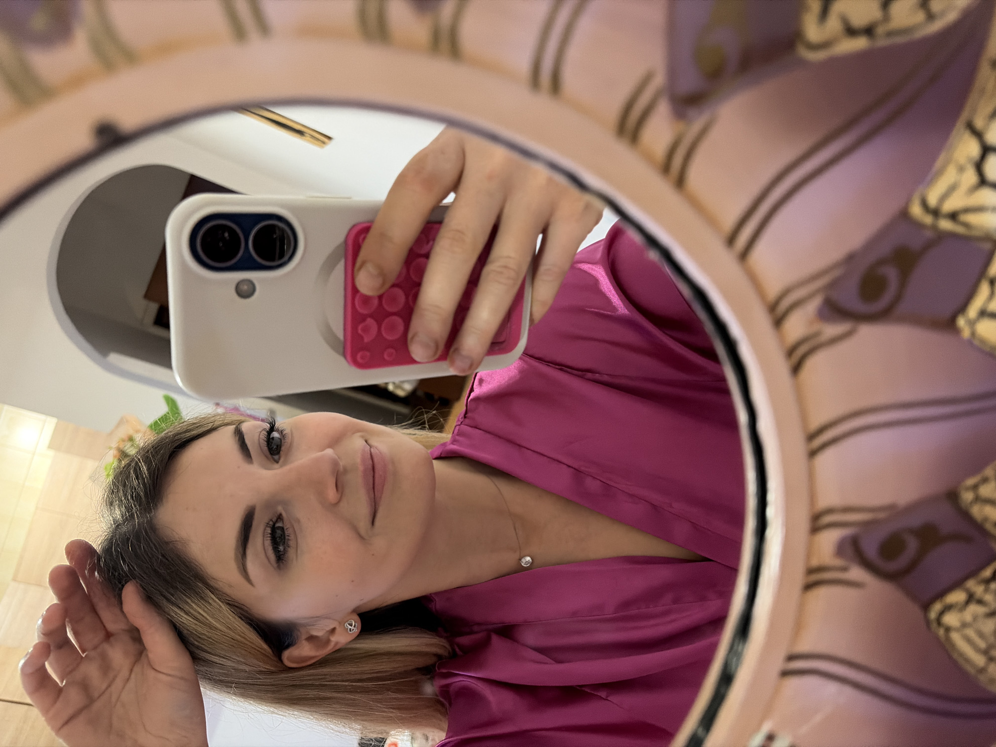 A young woman is reflected in a decorative mirror, taking a selfie with her smartphone. She has blonde hair and is wearing a bright pink satin robe and a small diamond necklace. Her hand is raised to her head, and she is smiling at the camera. The mirror has an ornate frame with floral motifs. In the reflection, a blurry background shows a wall with what appears to be some lighting fixtures. The overall scene suggests a private setting, likely a bedroom or dressing area, possibly in Budva, Montenegro, given the location context. The lighting appears to be artificial, possibly from indoor lighting or a ring light used for the selfie. The woman's expression is cheerful and relaxed. There is no visible text in the image.