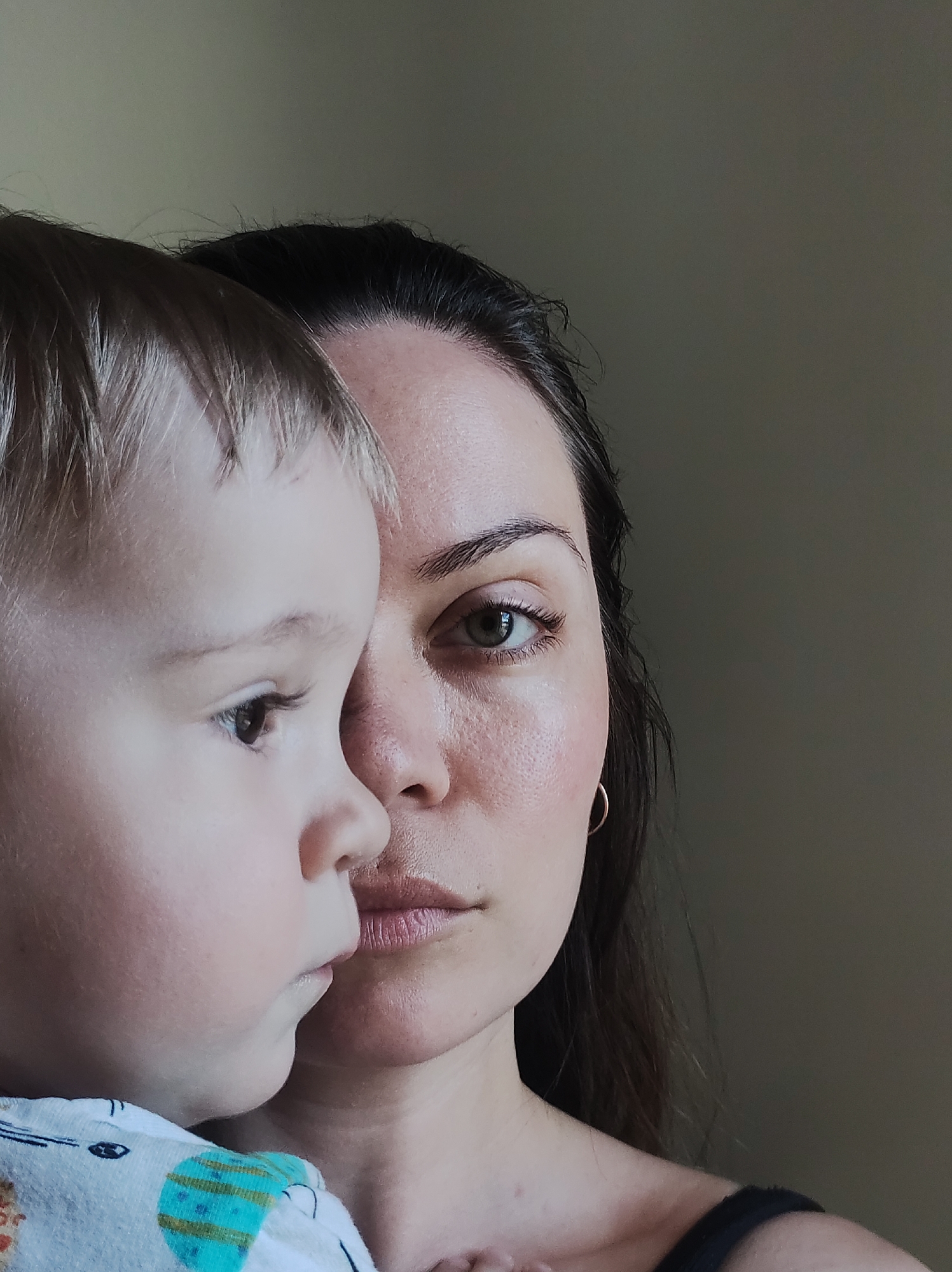 A medium close-up shot depicts a woman holding a young child. The woman is fair-skinned with dark hair pulled back from her face. Her left eye is visible, and she is wearing a small gold hoop earring in her left ear. The child has blonde hair and is wearing a patterned shirt. Both are looking to the left of the frame. The background is a solid, neutral color. The location is not discernible.