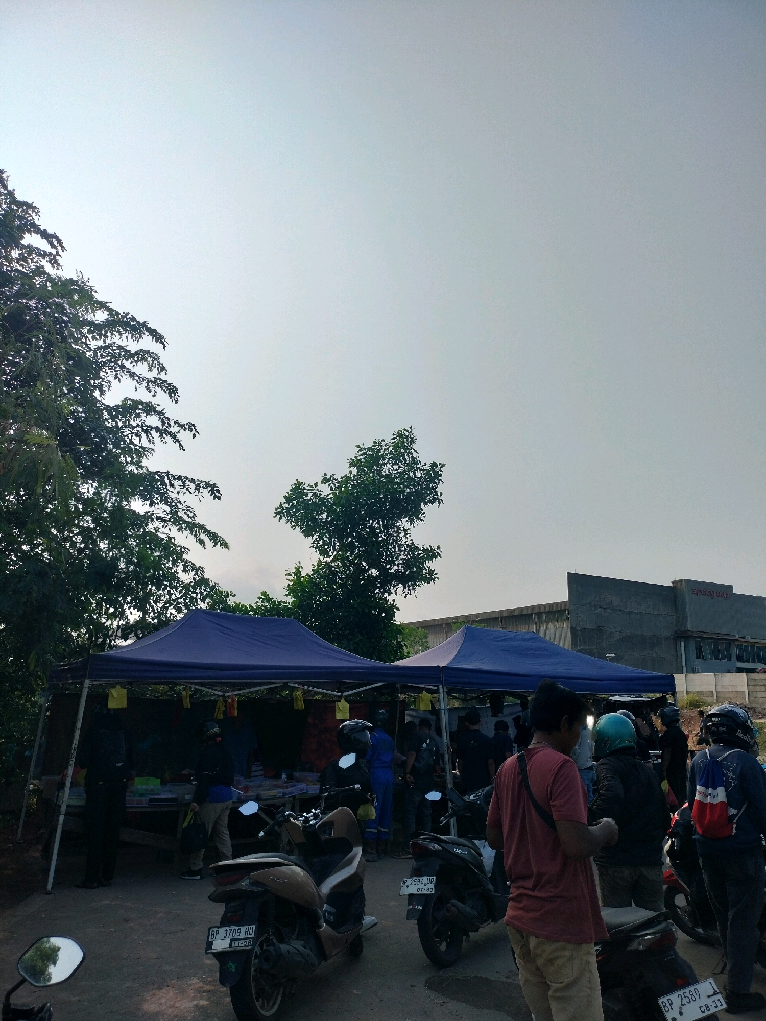 This is a medium-shot, eye-level, vertical, outdoor photograph taken during the day. The scene depicts a street market with two blue pop-up tents set up. In front of the tents, several motorcycles are parked. A group of people, some wearing helmets, are gathered around the tents, suggesting they are browsing or interacting with vendors. Some individuals appear to be customers, while others might be vendors. The background shows a building with signage, possibly an industrial facility, and trees lining the perimeter. The sky is overcast and bright. The overall impression is of a casual market or community gathering in a semi-urban or industrial area. It is likely located in the city of Batam, Indonesia.